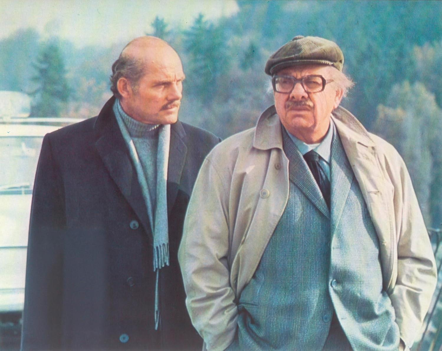 Zwei Männer in Mänteln stehen draußen. Einer hat eine Glatze und einen Schnurrbart, der andere trägt eine Mütze und eine Brille. Bäume sind im Hintergrund.