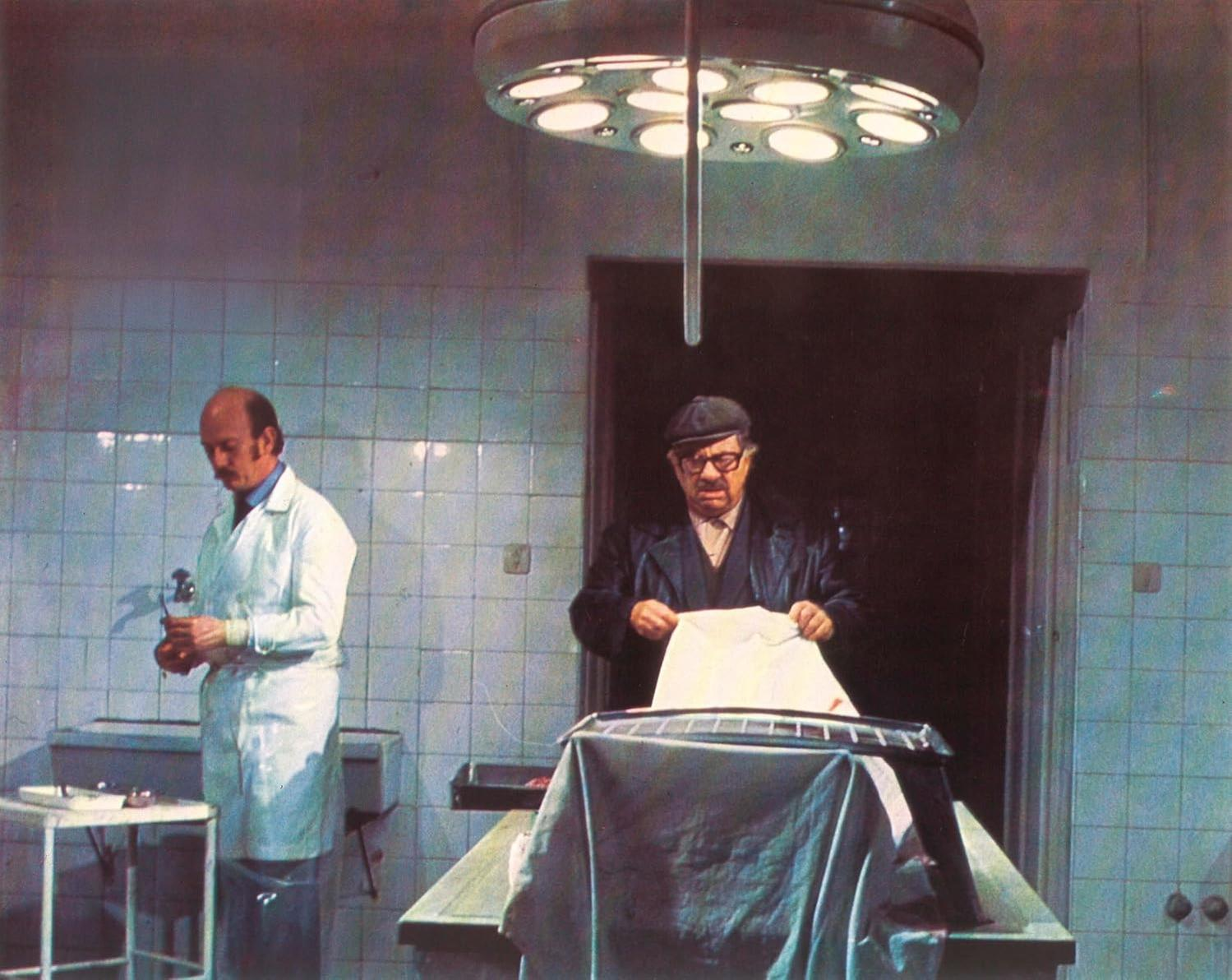 Zwei Männer in einem Operationssaal. Einer bereitet Instrumente vor, der andere untersucht ein Blatt. Helle Lichter hängen über einem Tisch.