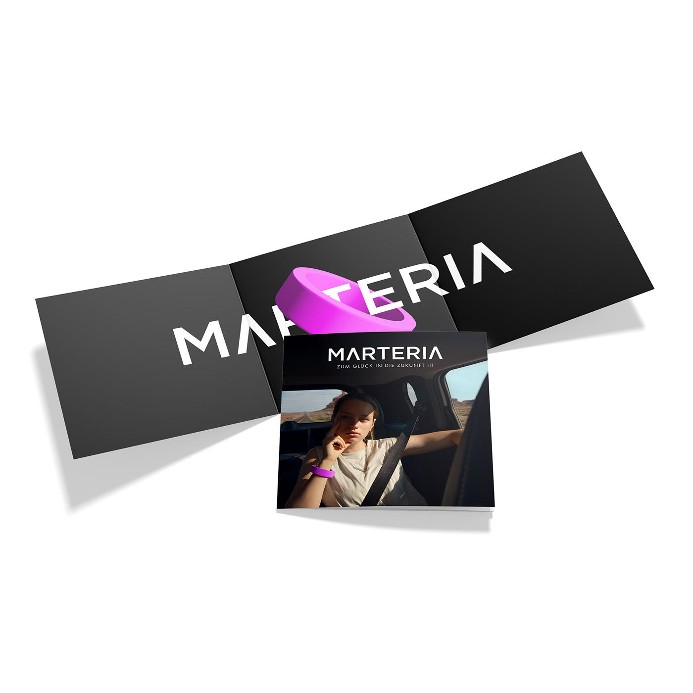 Das Bild zeigt eine Broschüre mit dem Wort "MARTERIA" und einer Frau in einem Auto. Es gibt einen pinken Ring und schwarze Karten.