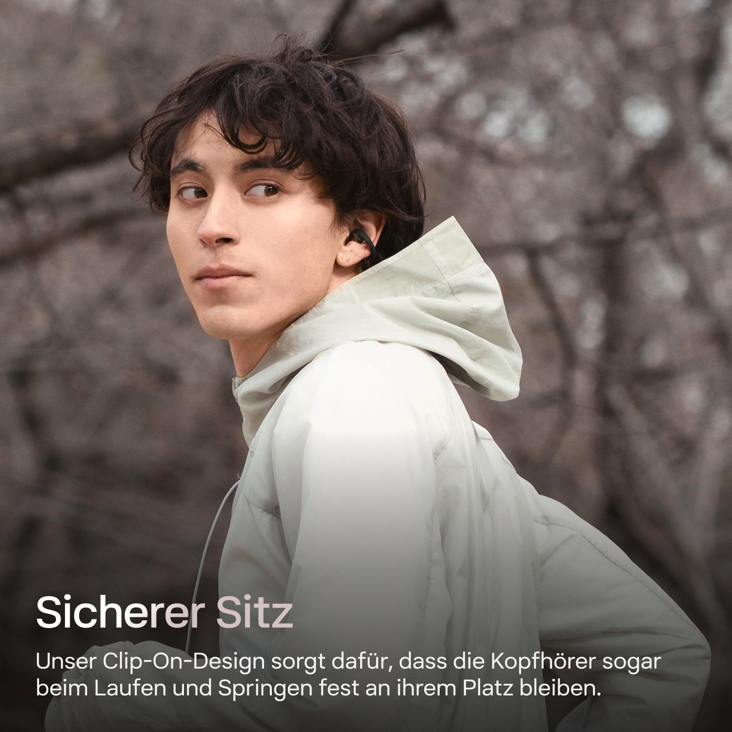 Ein junger Mann mit einer leichten Jacke und Ohrhörern, der nach rechts blickt. Der Hintergrund zeigt verschwommene Bäume.