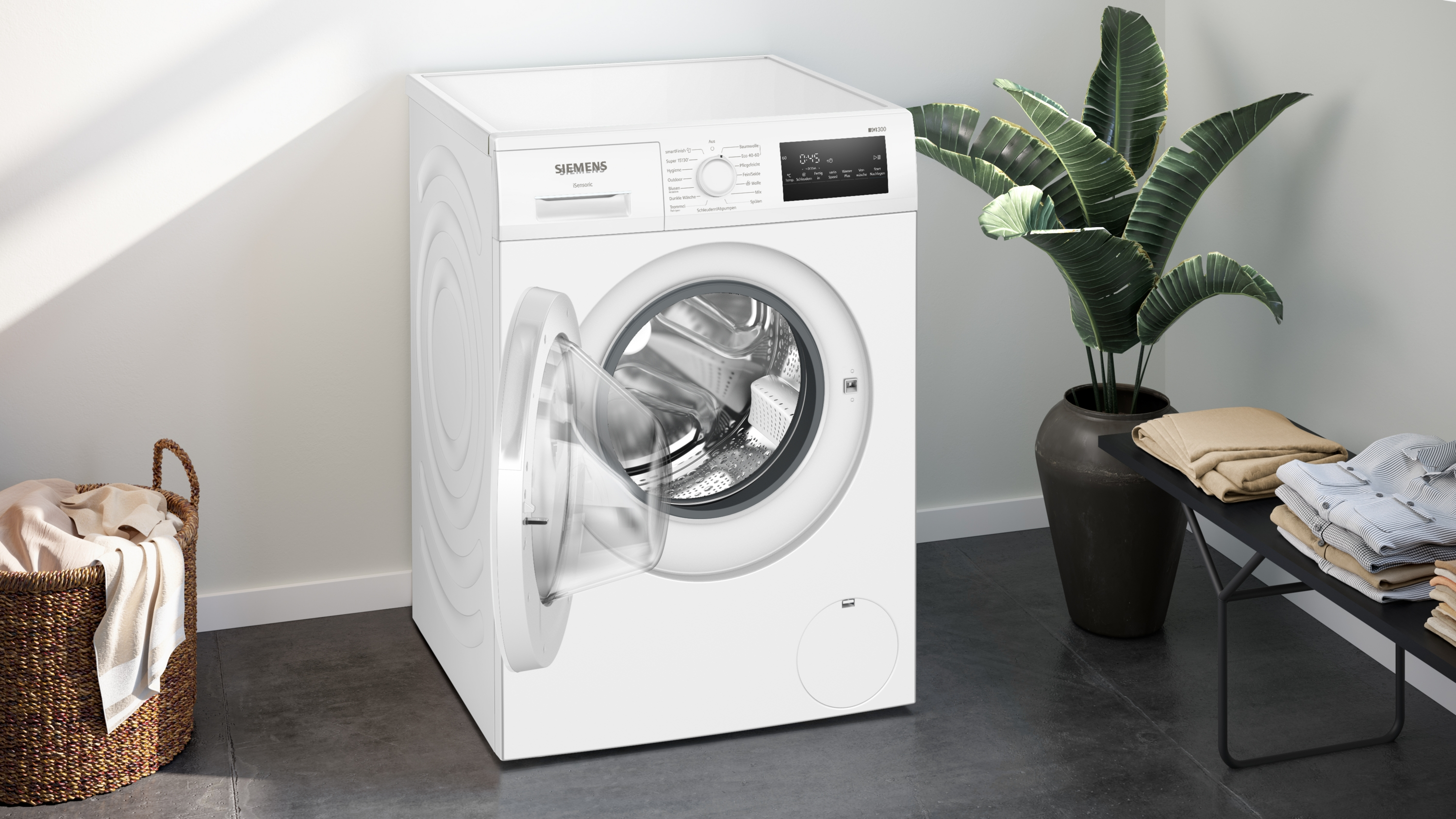 Eine weiße Waschmaschine ist geöffnet, neben einer Pflanze im Topf und einem Korb mit Kleidung.