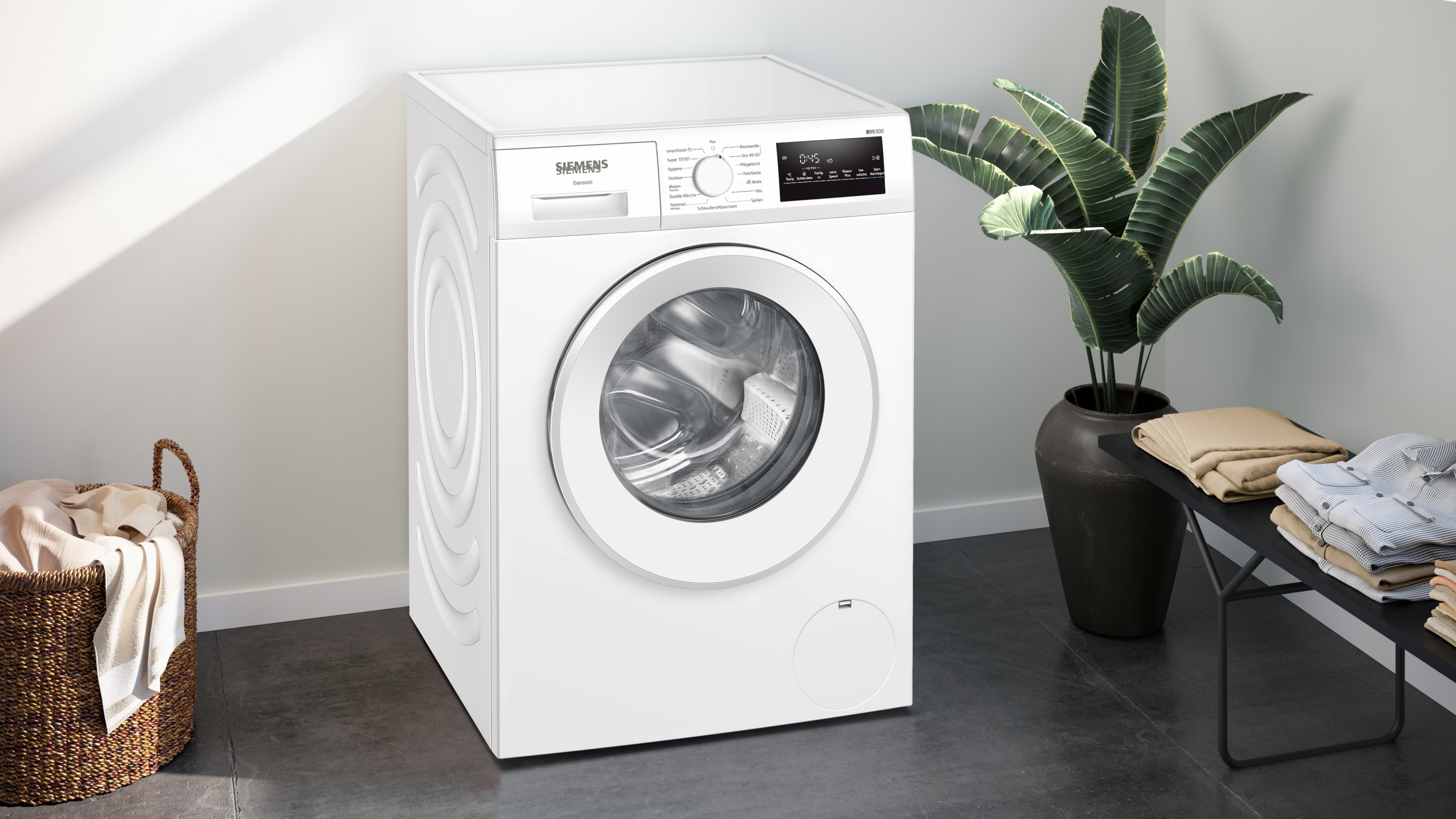 Eine weiße Siemens-Waschmaschine steht neben einer Pflanze. Links ein Wäschekorb und rechts Kleidung auf einem schwarzen Tisch.