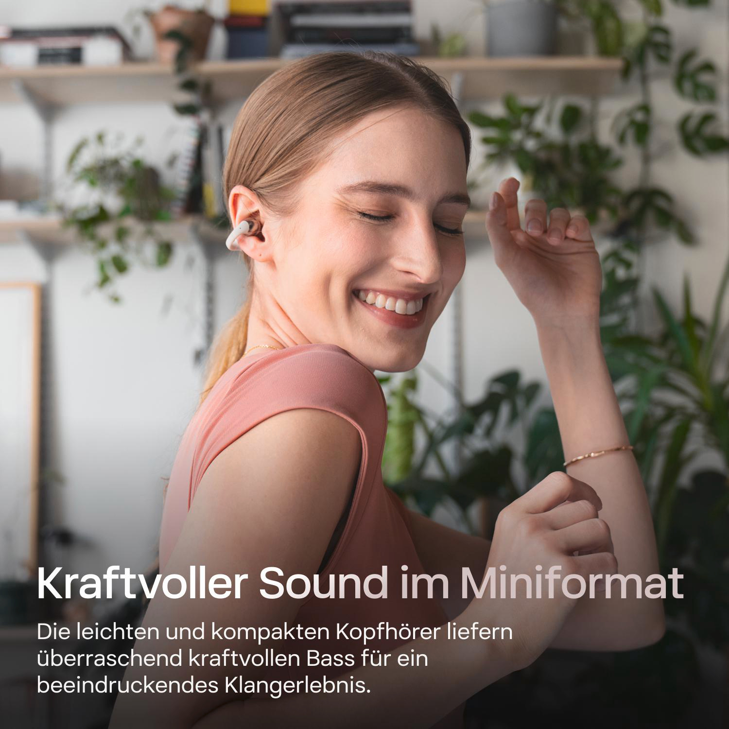 Eine lächelnde Frau mit Ohrhörern, die eine Hand hebt. Im Hintergrund ist ein Regal mit Pflanzen zu sehen.