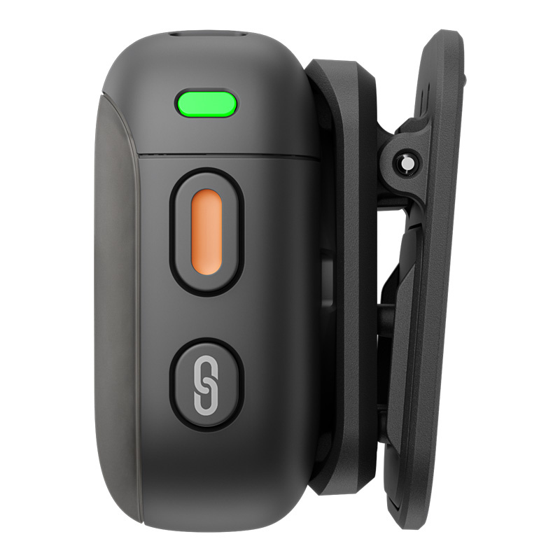Czarne urządzenie z zielonym światłem, pomarańczowym przyciskiem i przyciskiem 'S', przypięte do czarnego uchwytu. Na białym tle.