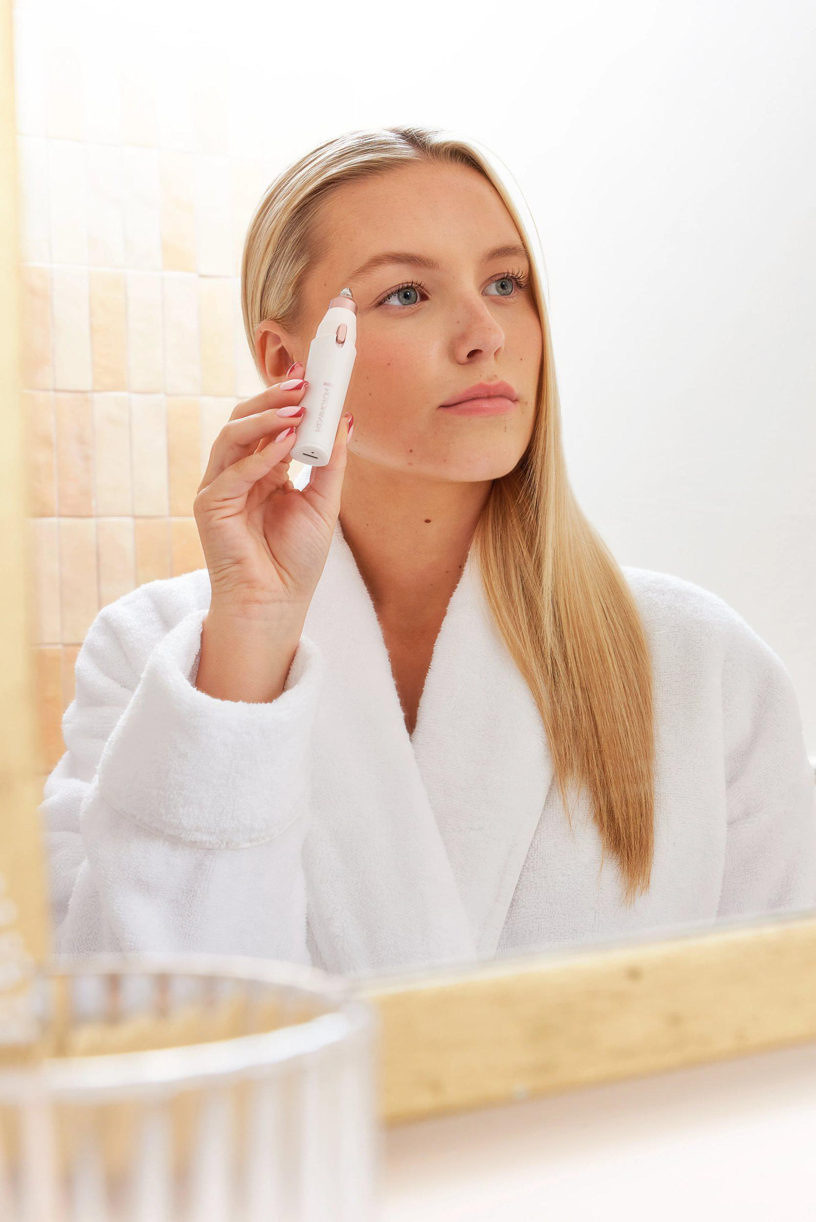 Blonde Frau im weißen Bademantel benutzt ein Gesichtsgerät. Sie schaut in den Spiegel. Beige geflieste Wand im Hintergrund.