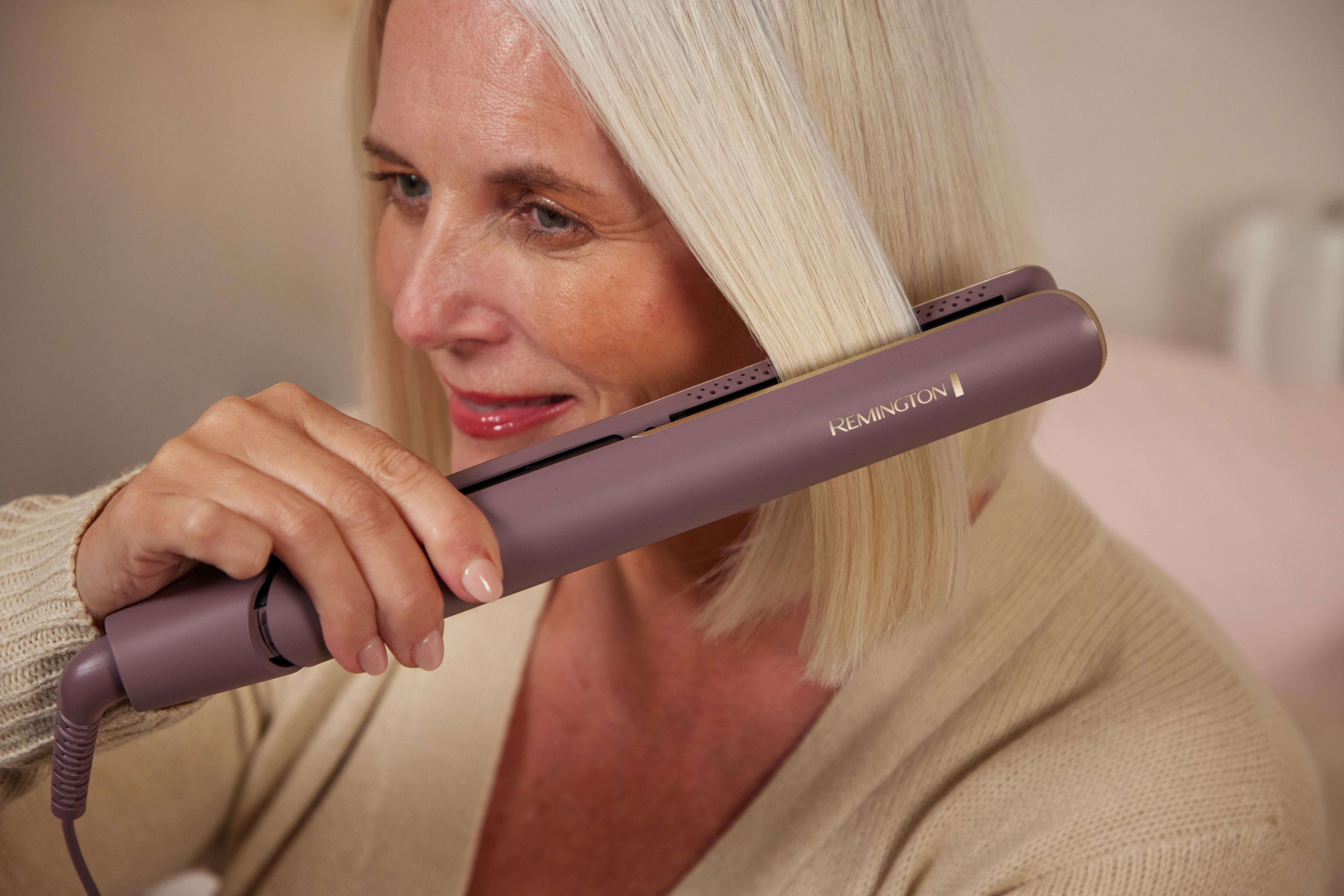 Eine Frau mit blonden Haaren benutzt ein lila Remington Glätteisen, sie trägt einen beigefarbenen Pullover. Der Hintergrund ist unscharf.