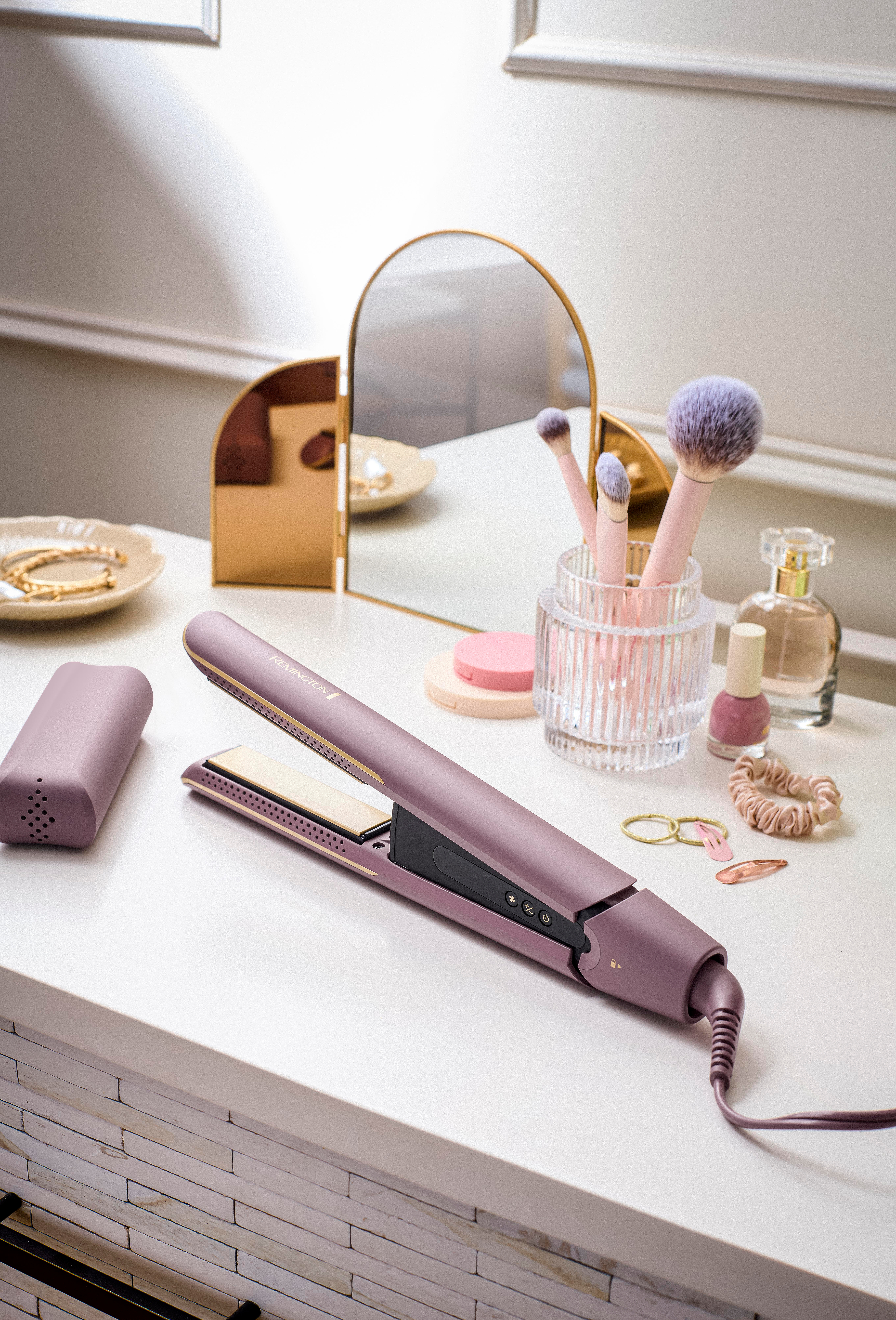 Ein Glätteisen, Make-up-Pinsel und Accessoires liegen auf einer weißen Oberfläche. Ein Spiegel und Parfüm sind im Hintergrund.