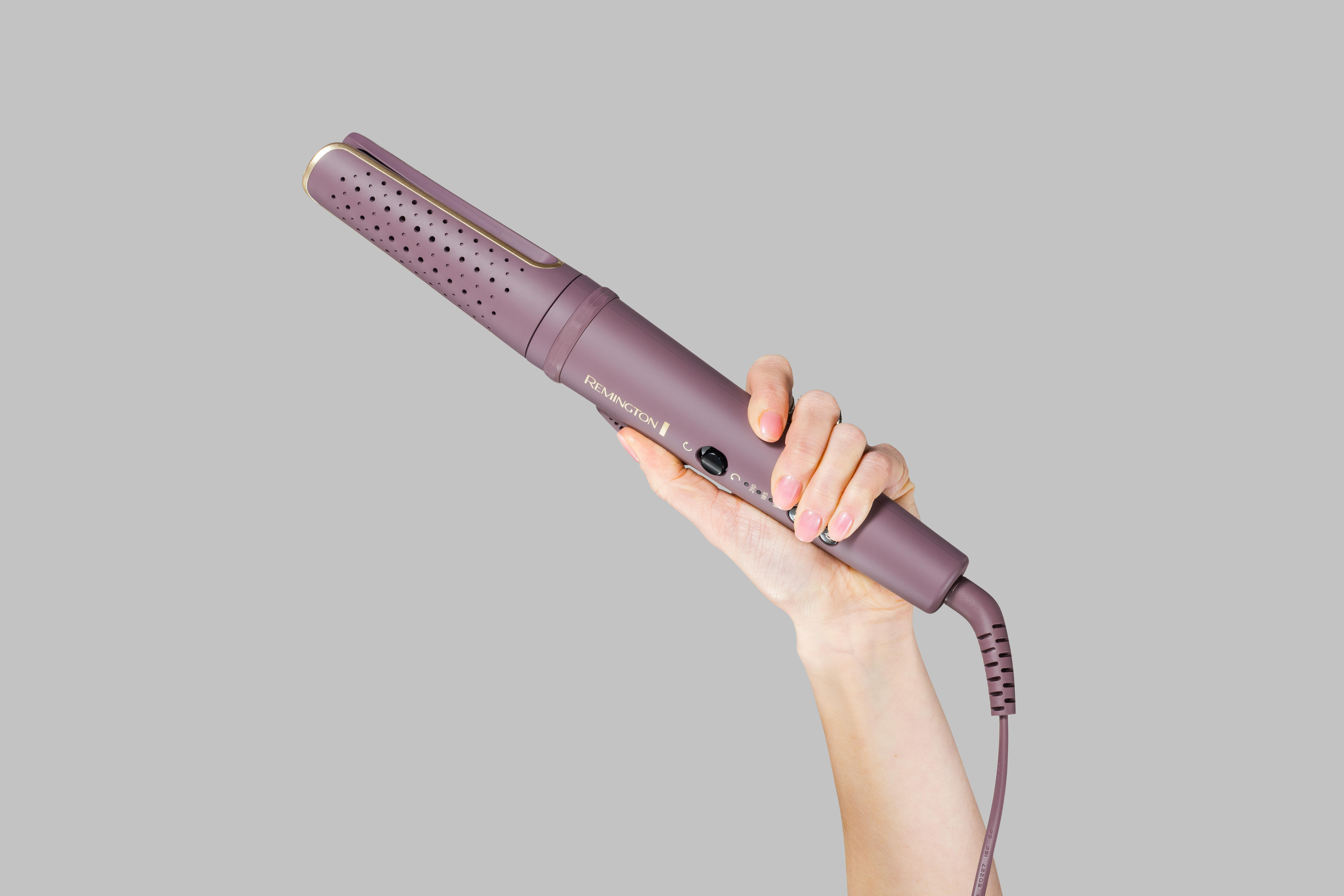 Eine Hand hält ein lila Haarstyling-Werkzeug vor grauem Hintergrund. Das Werkzeug hat ein Kabel und der Markenname ist sichtbar.