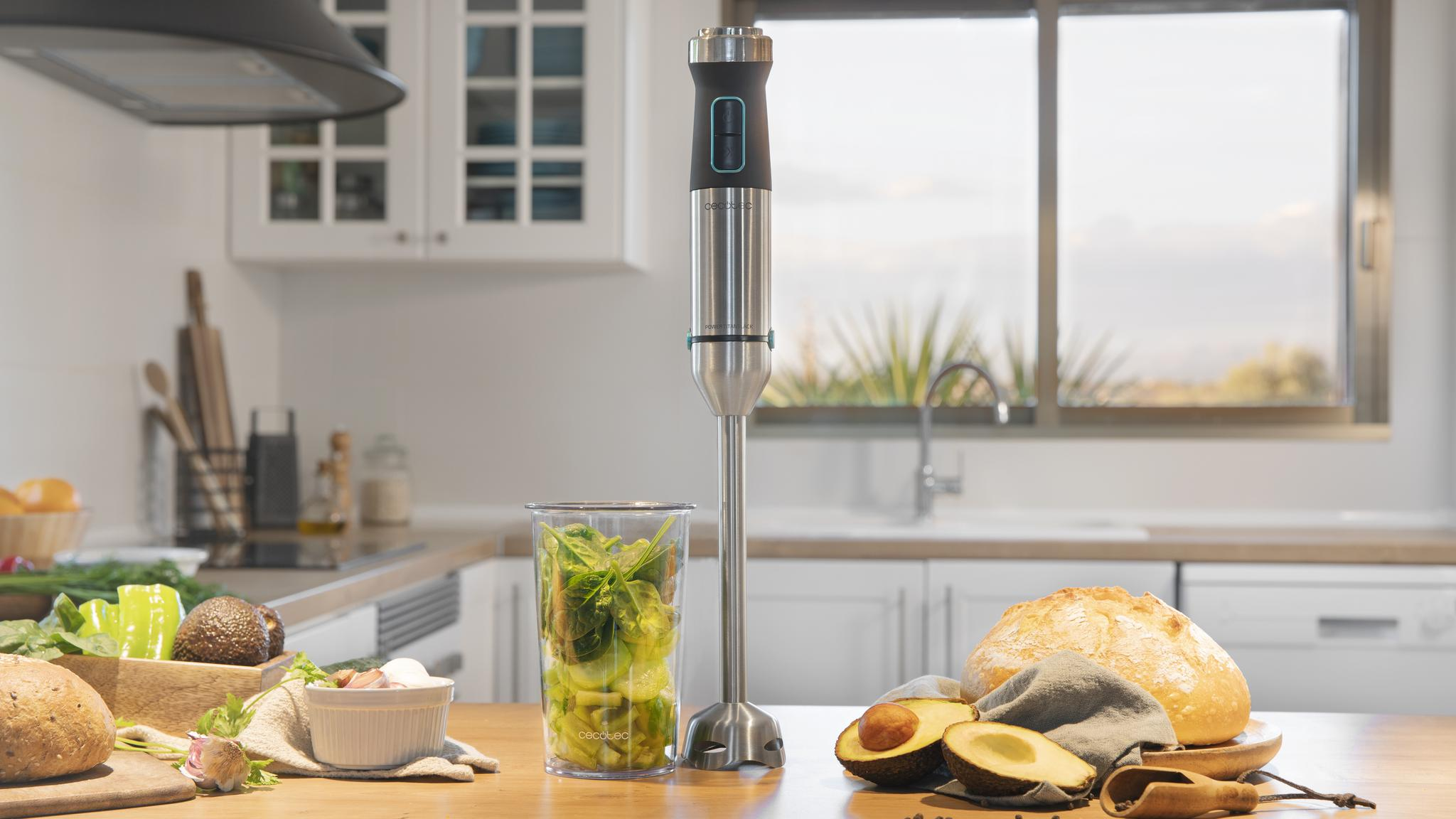 Una escena de cocina con una batidora, aguacate, pan y verduras sobre una mesa de madera. Una ventana y armarios en el fondo.
