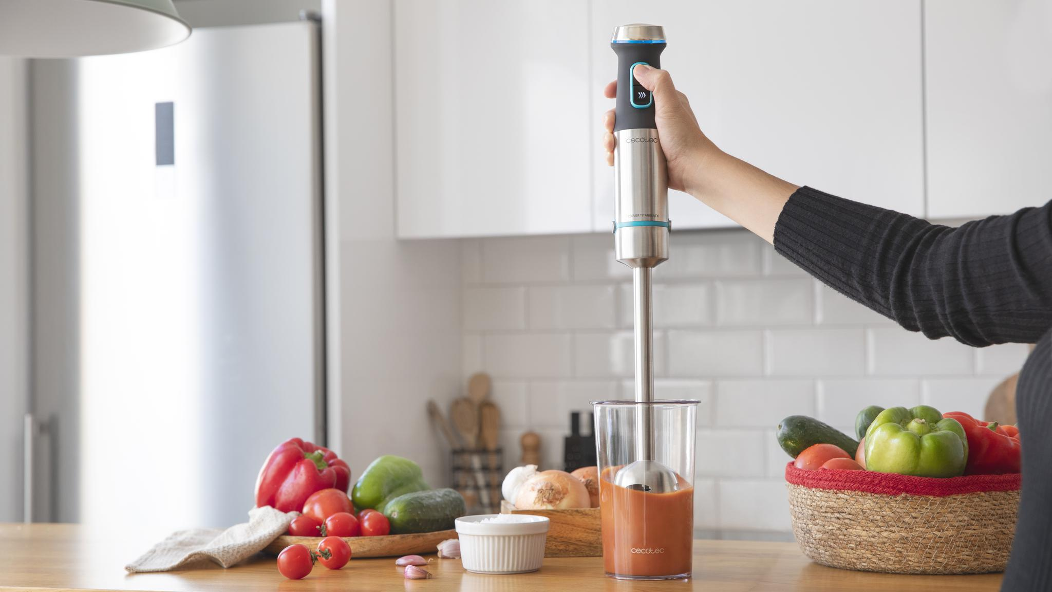 Una persona usa una batidora de mano en una cocina. En la mesa hay verduras como tomates, pimientos y calabacines.