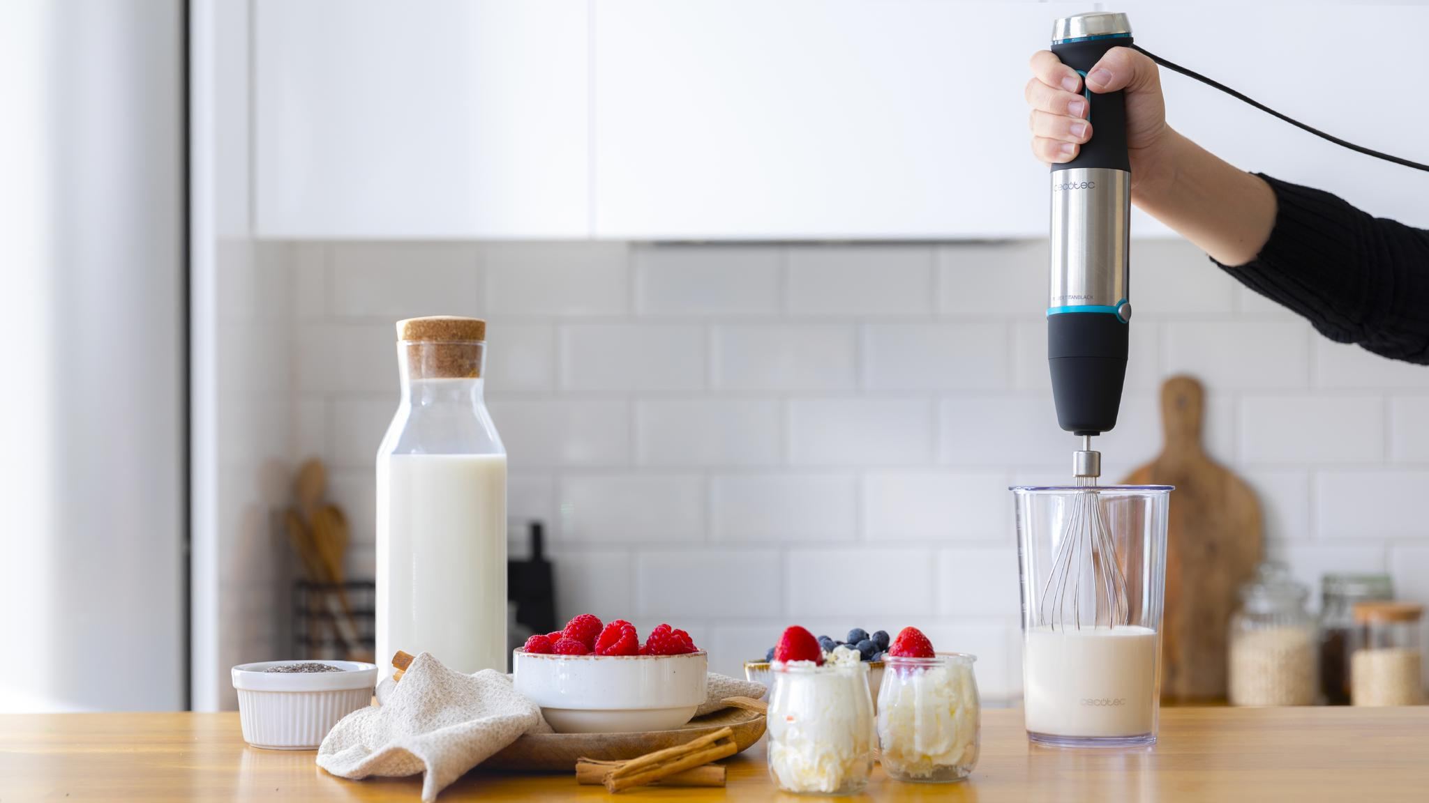 Mano sosteniendo una batidora sobre un vaso con leche. Cuencos con bayas, botella de leche y otros ingredientes en mesa de madera.