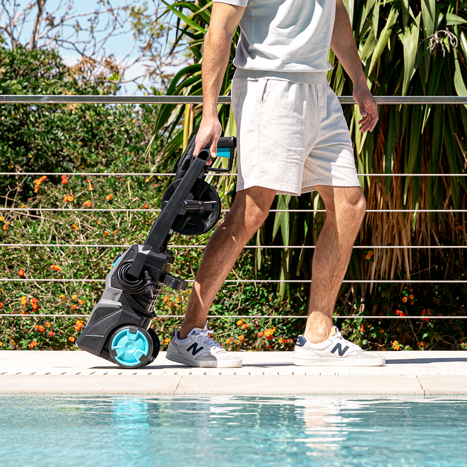 Un hombre camina, llevando una hidrolavadora. Usa pantalones cortos y zapatillas. Una piscina y vegetación al fondo.