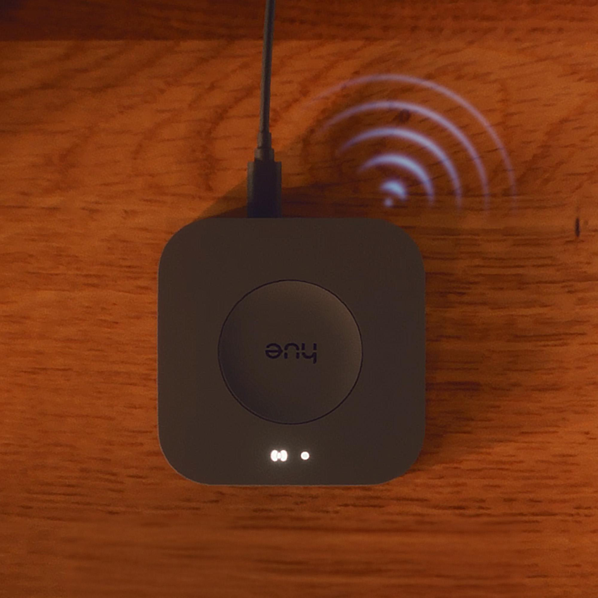 Ein dunkelgrauer Philips Hue Bridge liegt auf Holz. Ein Kabel verbindet es mit einer unsichtbaren Quelle. Drahtlose Signalbögen darüber.