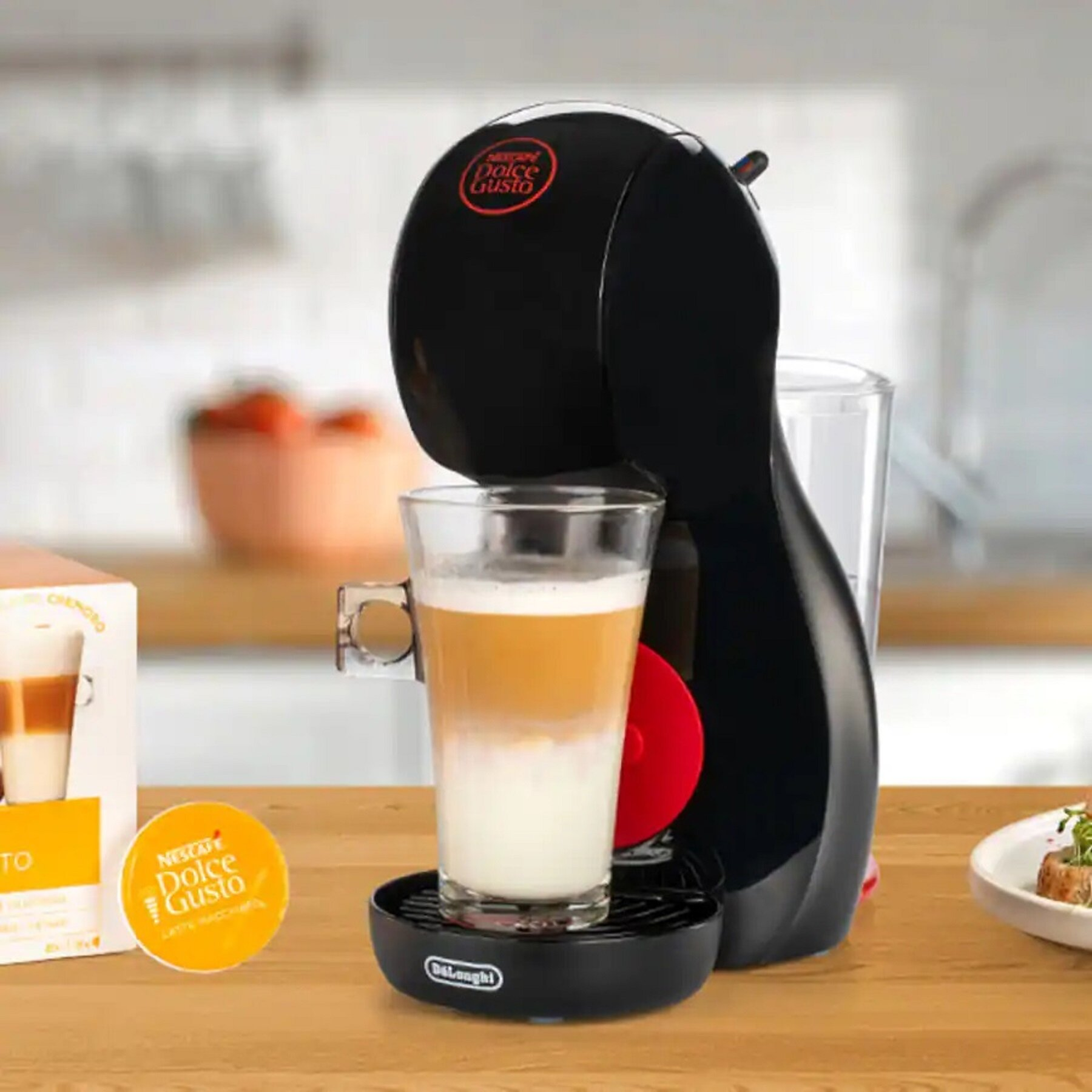 Czarny ekspres do kawy Dolce Gusto z latte w szklance. Kapsułka i pudełko leżą na drewnianym stole.