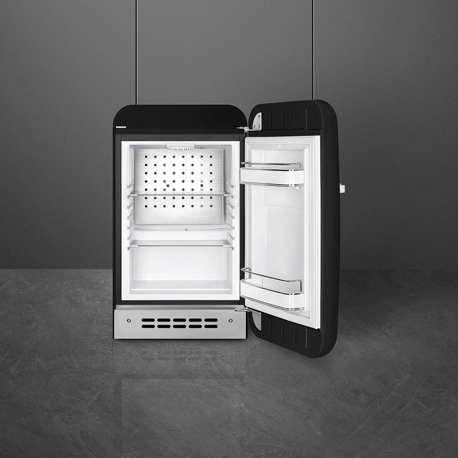 Mini frigo nero in stile retrò con la porta aperta. Interno bianco, con ripiani. Sfondo e pavimento grigi.