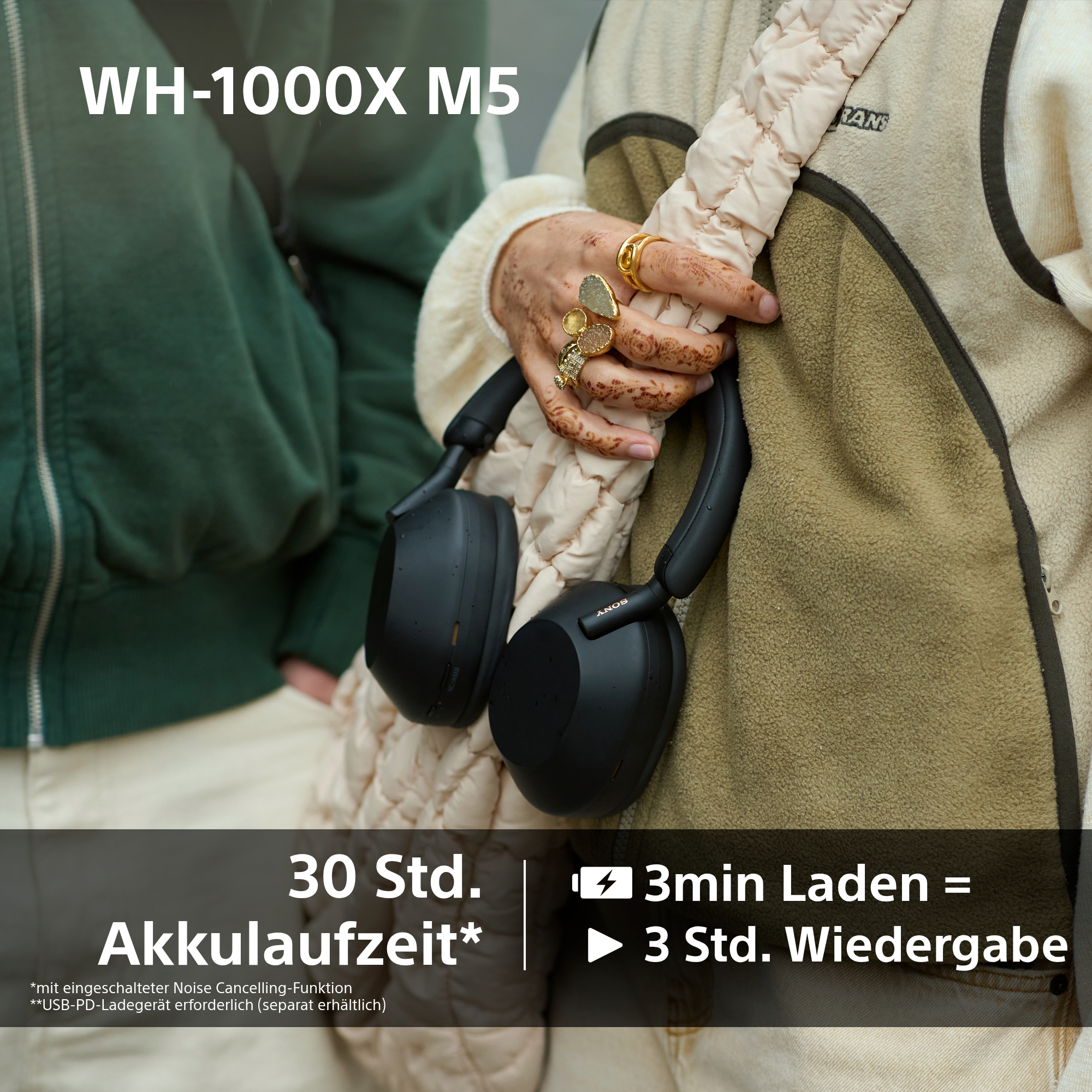 Eine Person hält schwarze Sony-Kopfhörer. Die Person trägt Ringe und eine beige Jacke, der Hintergrund ist grün.