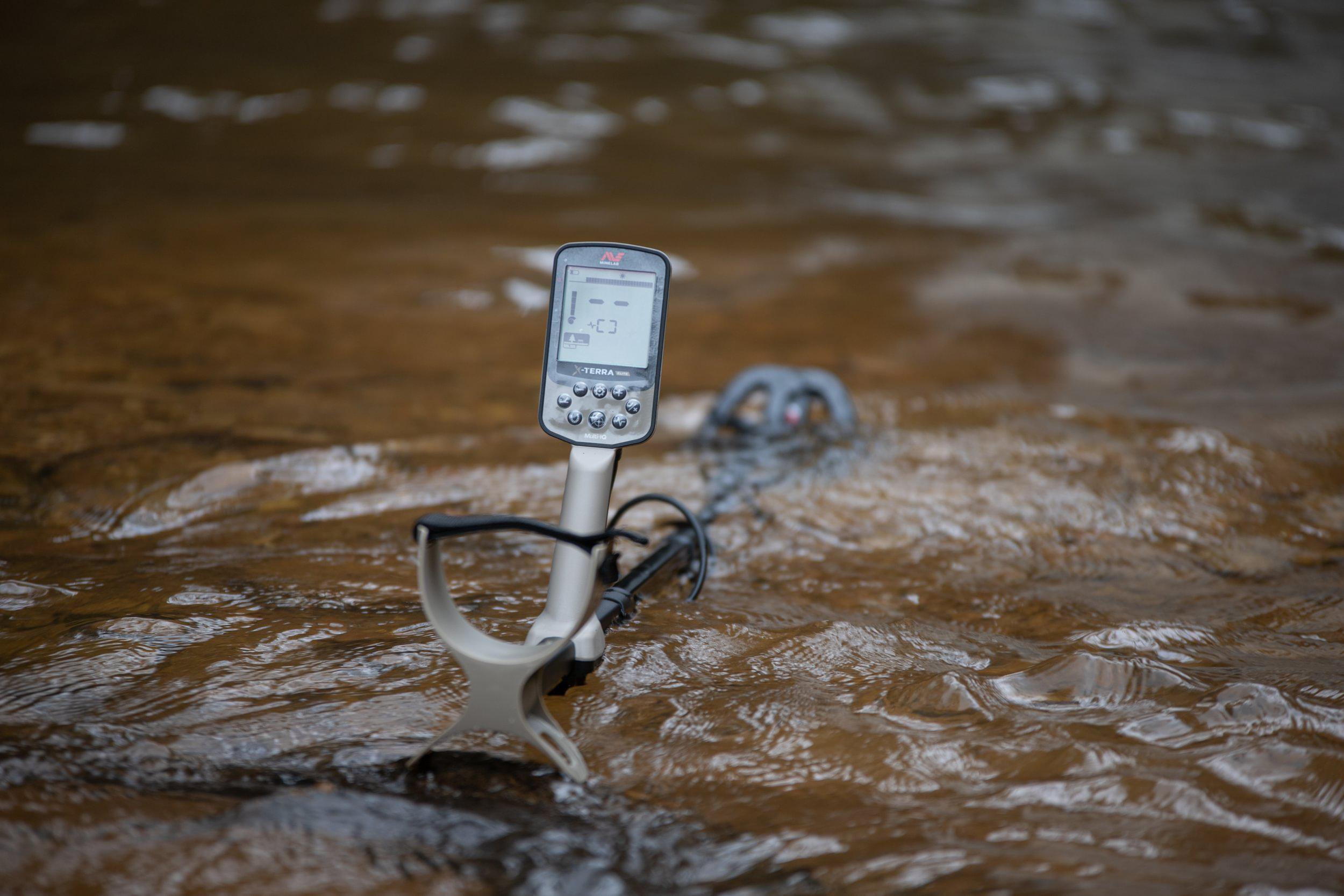 Ein Metalldetektor ist in einem Bach untergetaucht. Der Bildschirm zeigt eine Anzeige. Das Wasser ist braun. Der Detektor ist silber und schwarz.