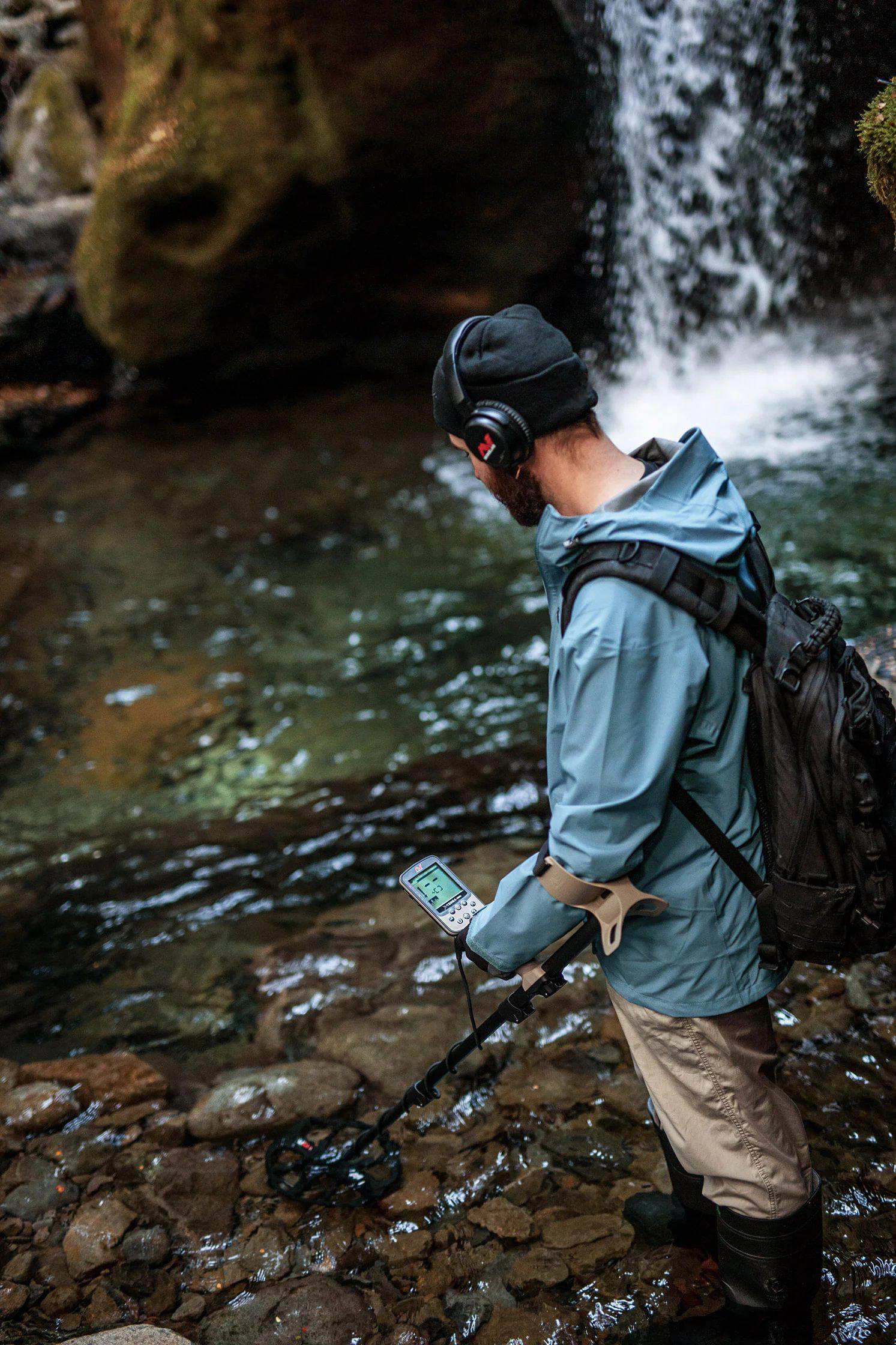 Ein Mann mit Metalldetektor steht in einem Bach in der Nähe eines Wasserfalls. Er trägt einen schwarzen Hut, eine blaue Jacke und Stiefel.