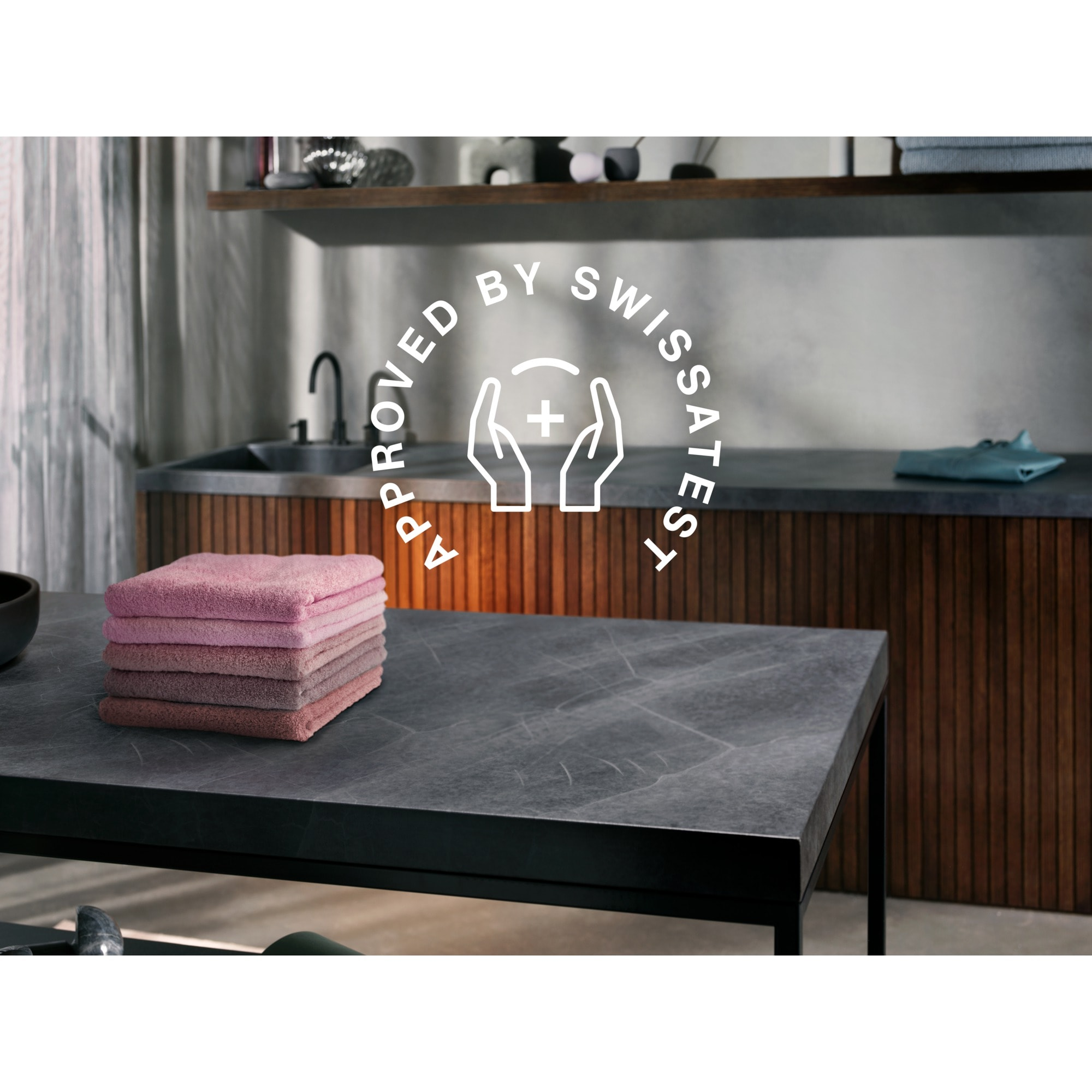 Pila de toallas rosas sobre una mesa negra. El fondo muestra un lavabo, paneles de madera y un logo 'Approved by Swissatest'.