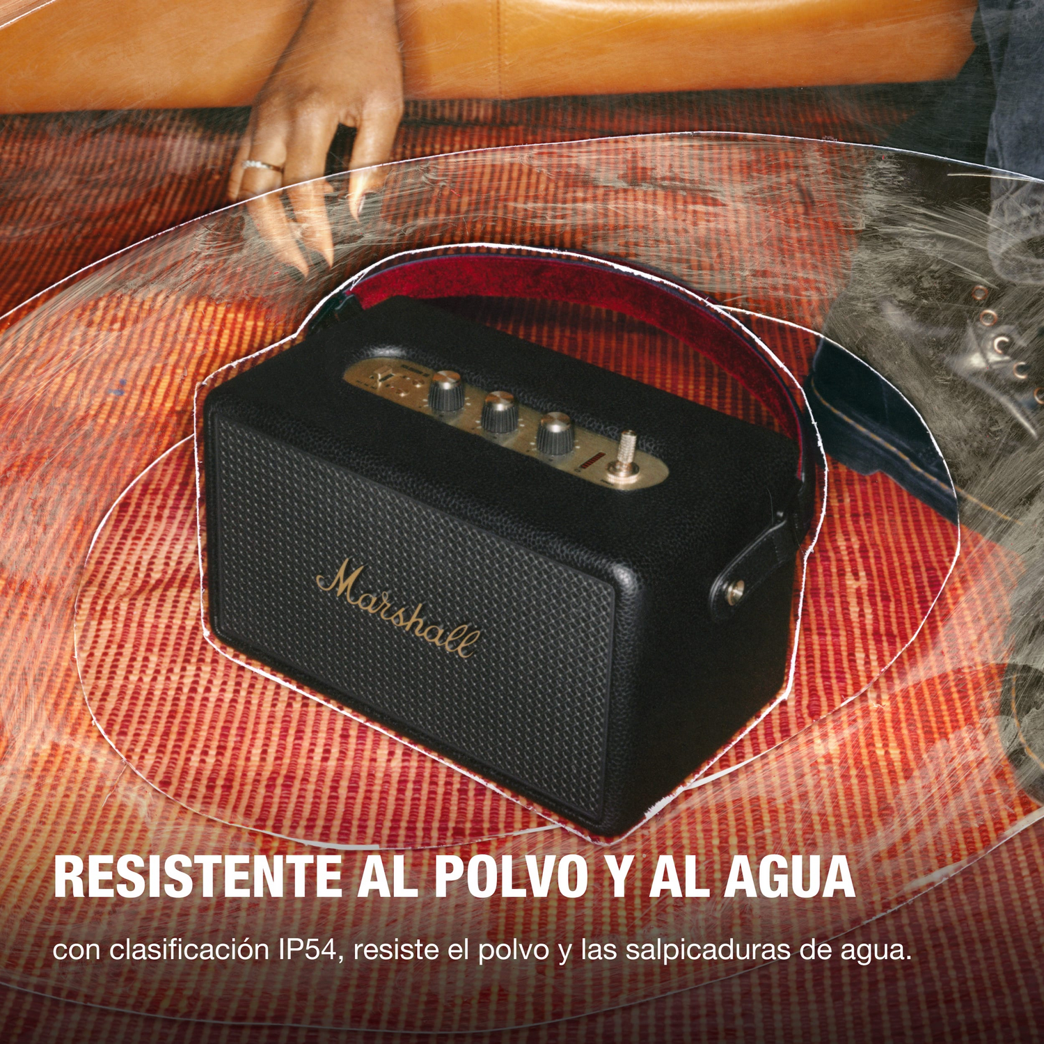Altavoz Marshall negro sobre superficie roja estampada. Mano, anillo y texto 'RESISTENTE AL POLVO Y AL AGUA' visibles.