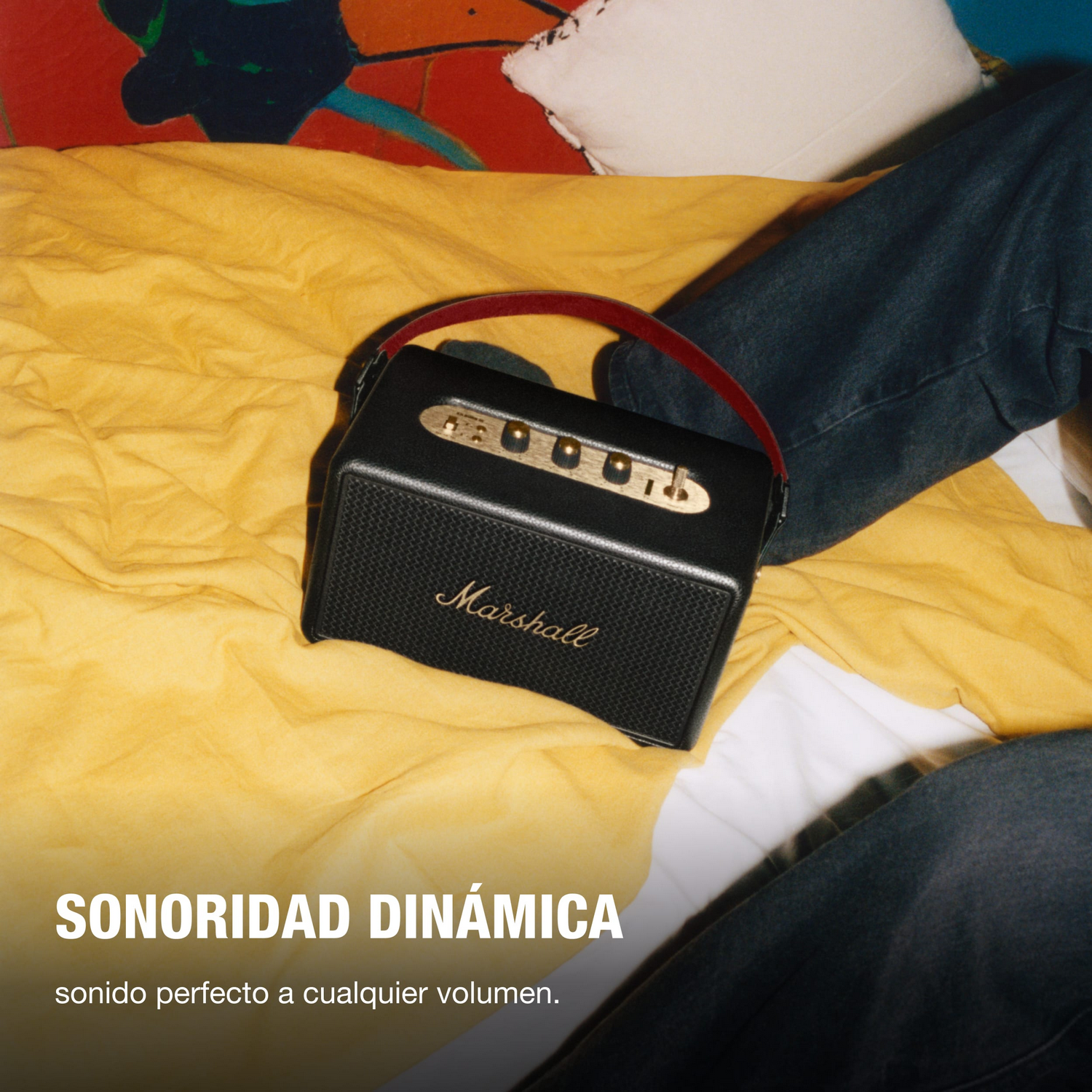 Altavoz Marshall negro con asa roja sobre manta amarilla, vaqueros en el fondo. Texto: Sonido dinámico. Sonido perfecto a cualquier volumen.