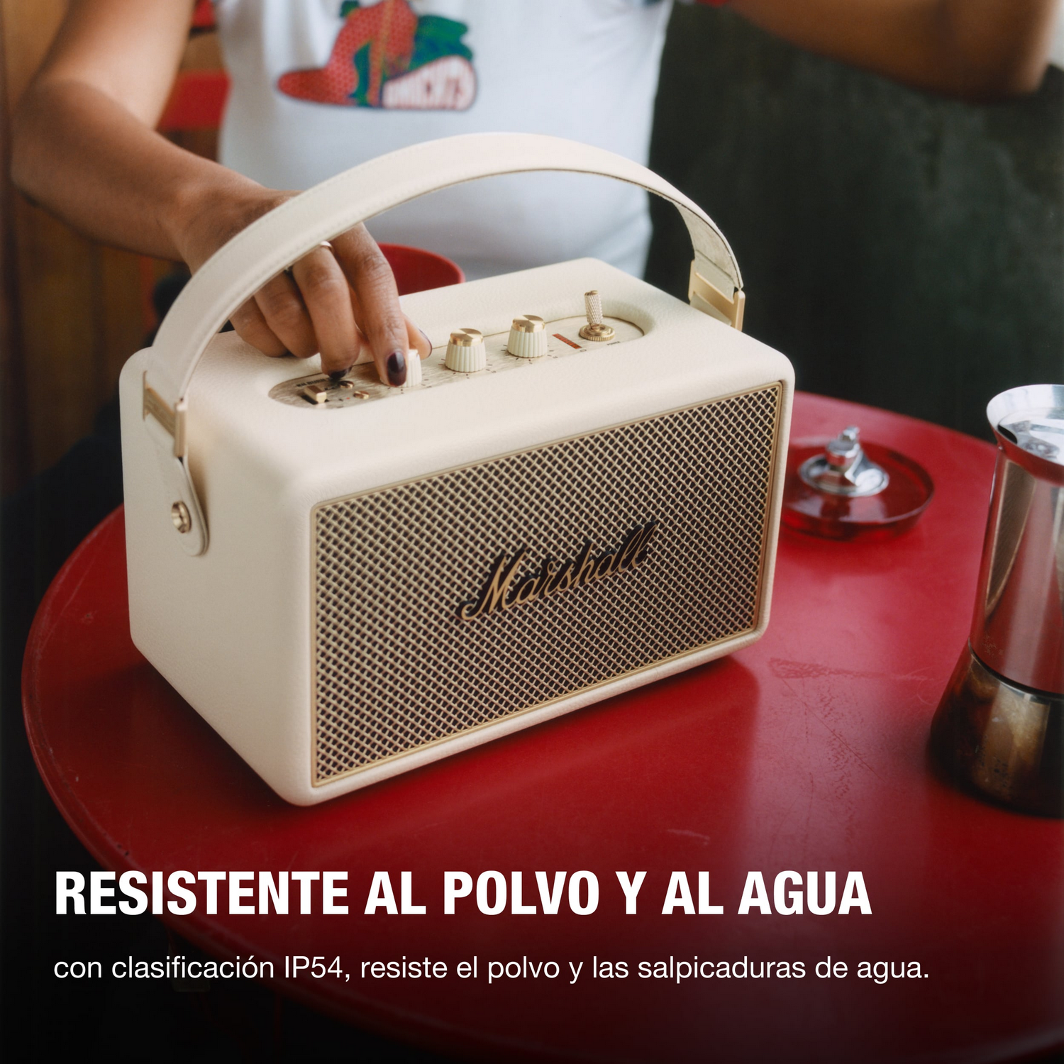 Una mano ajusta un altavoz Marshall crema en una mesa roja. El altavoz tiene una malla y un asa doradas.