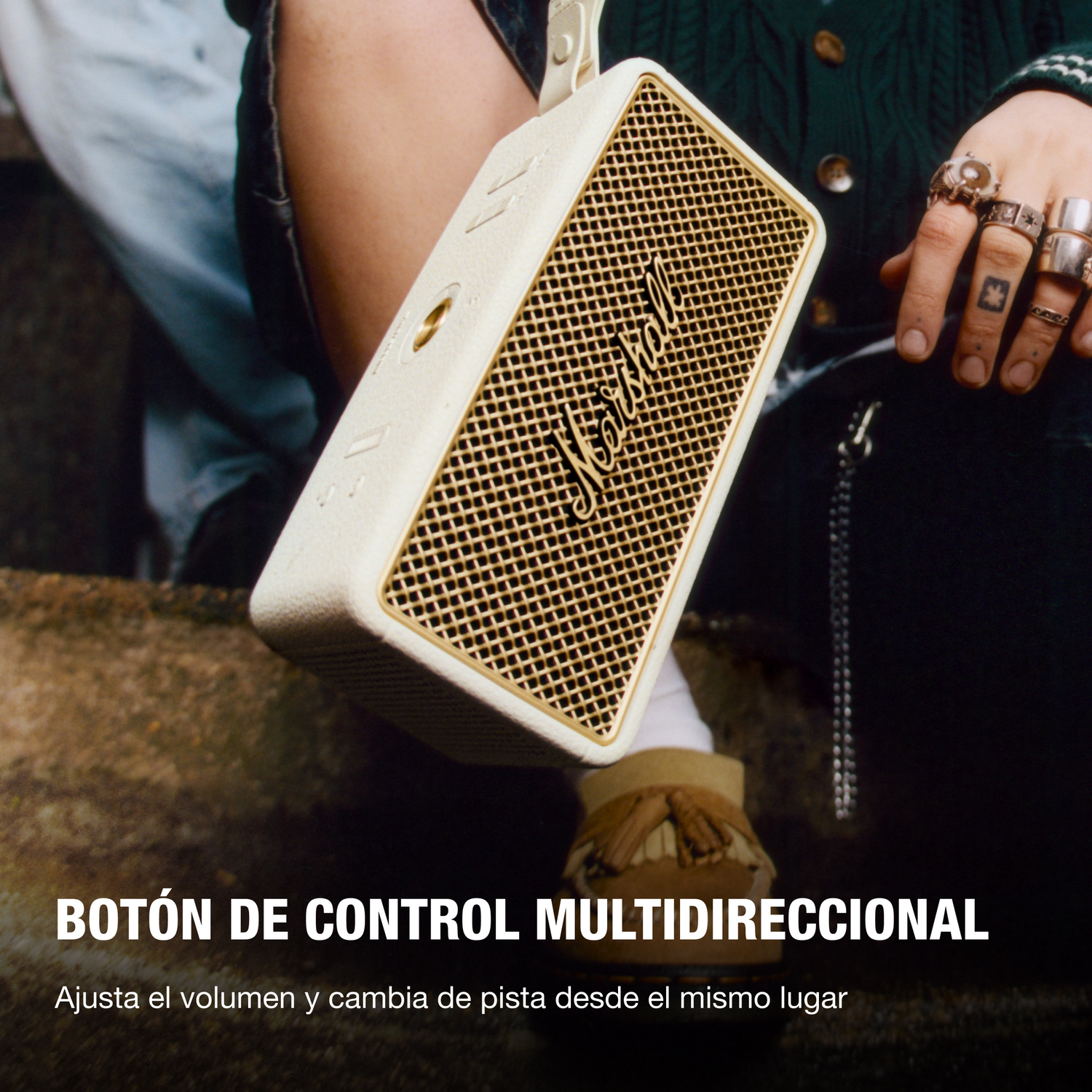 Una persona sostiene un altavoz Marshall blanco y dorado. La persona lleva anillos y un suéter verde. Abajo pone 'Botón de control multidireccional'.