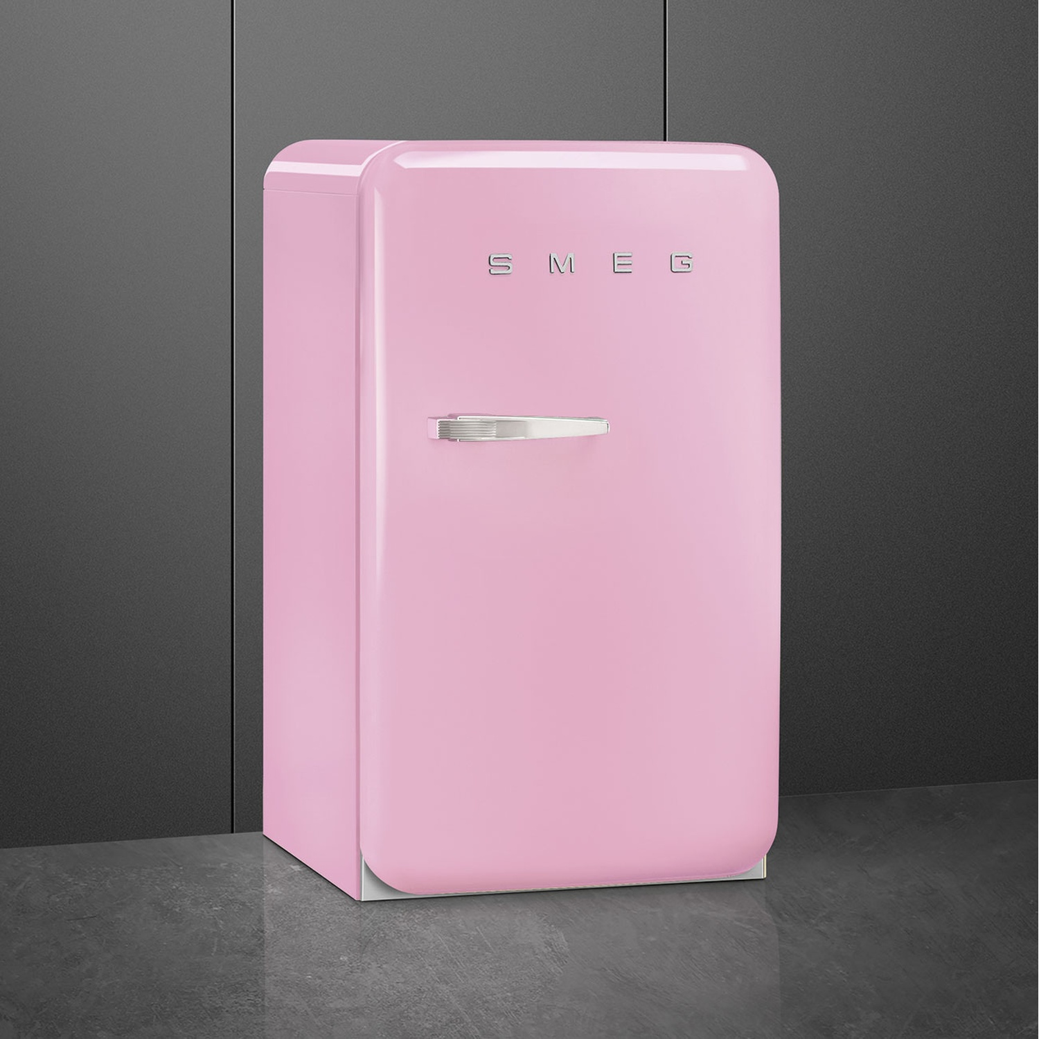 Un frigorifero SMEG rosa. Ha una maniglia argentata e il logo SMEG. È su un pavimento grigio scuro con uno sfondo grigio scuro.