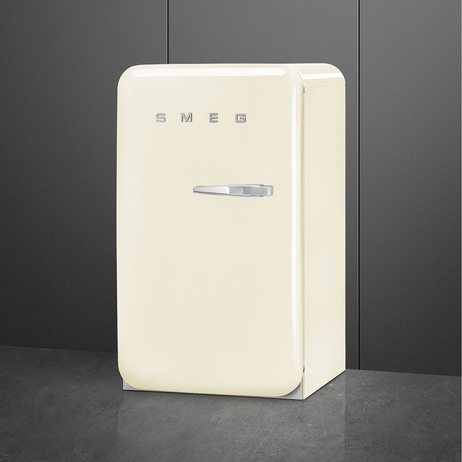 Un frigorifero Smeg color crema si trova contro un muro grigio scuro. Ha una maniglia argentata e il logo del marchio in alto.