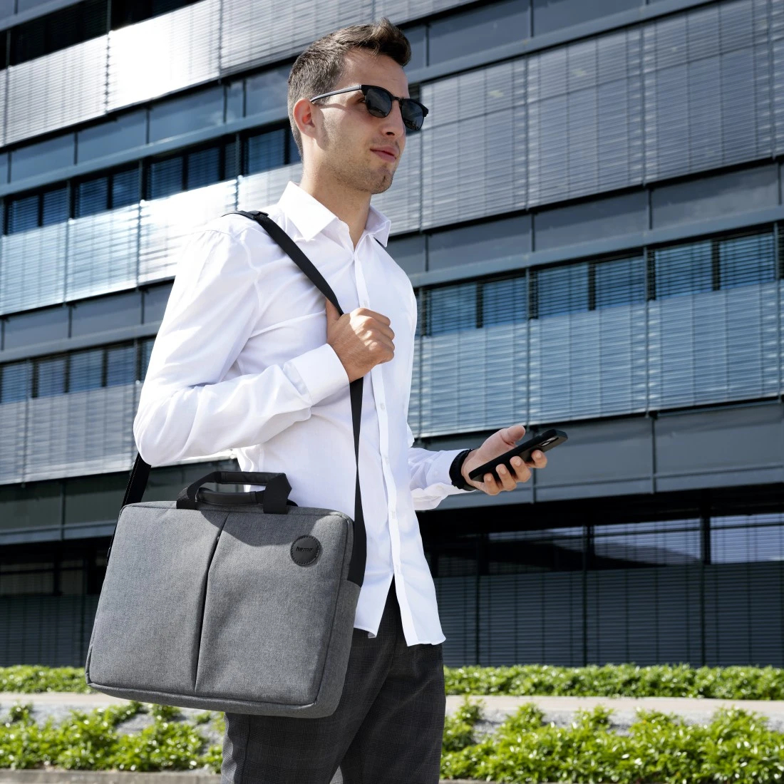 Un hombre con gafas de sol camina, lleva un maletín gris para portátil y sostiene un teléfono. Un edificio moderno y arbustos en el fondo.
