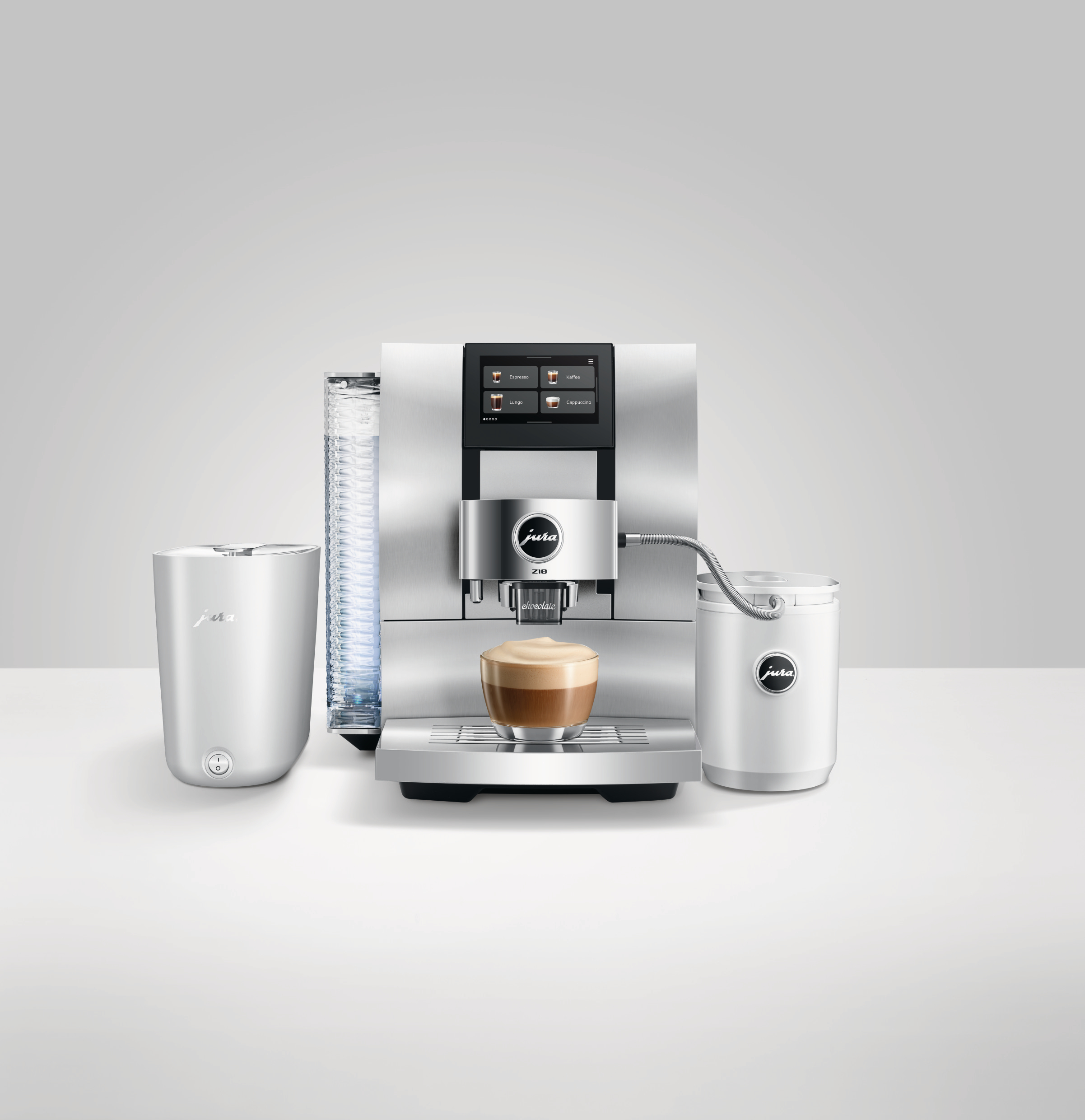 Eine silberne Jura-Kaffeemaschine mit Touchscreen, ein Glas Cappuccino und zwei weiße Behälter auf weißer Oberfläche.