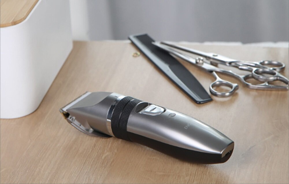 Srebrna maszynka do włosów, czarny grzebień i nożyczki na drewnianej powierzchni. Białe pudełko po lewej.