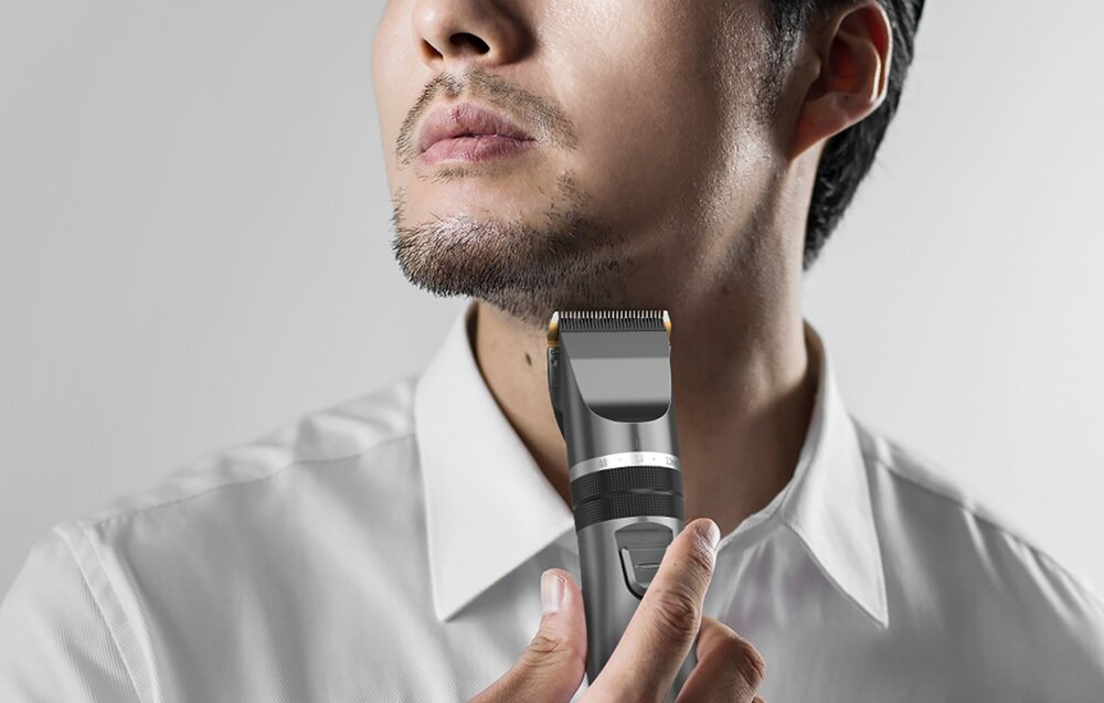 Mężczyzna goli brodę trymerem elektrycznym. Ma na sobie białą koszulę. Tło jest białe.