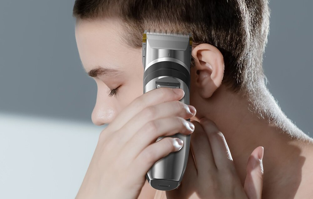 Osoba goli włosy maszynką elektryczną. Oczy osoby są zamknięte. Tło jest jasnoszare.