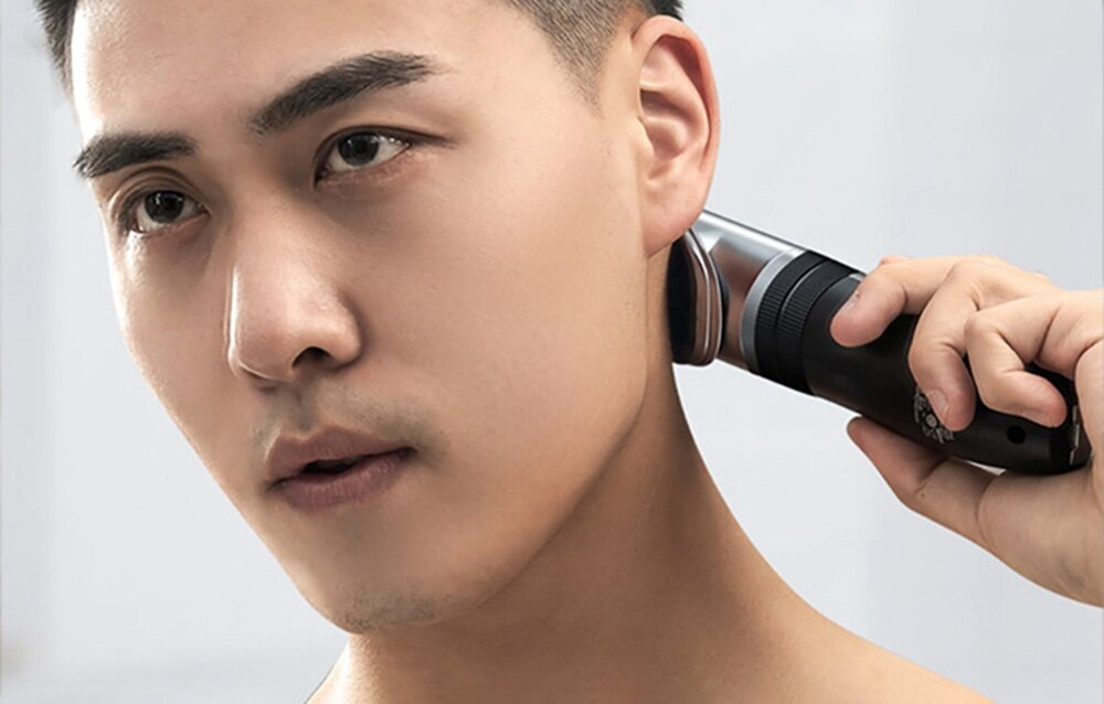 Mężczyzna goli szyję maszynką elektryczną. Ma ciemne włosy i trzyma maszynkę przy szyi. Tło jest białe.