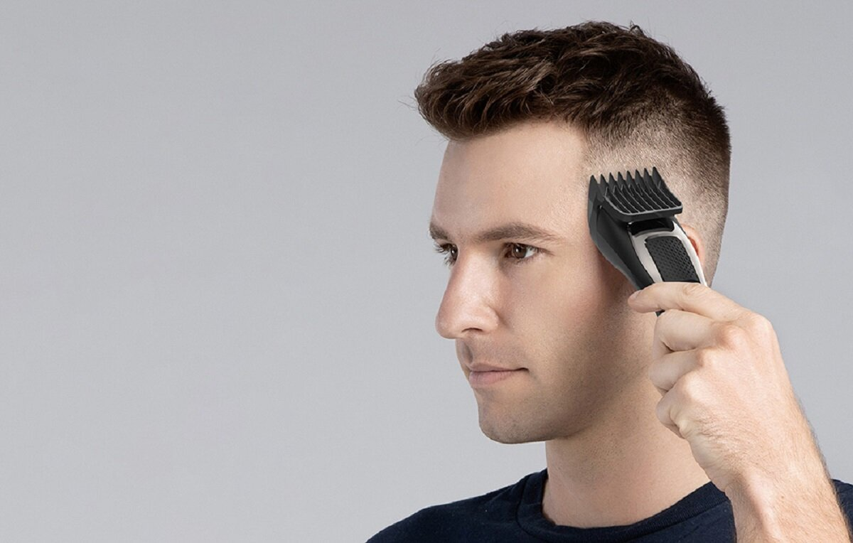 Mężczyzna przycina włosy czarno-srebrną elektryczną maszynką do strzyżenia. Ma na sobie ciemnoniebieską koszulę i krótkie brązowe włosy.