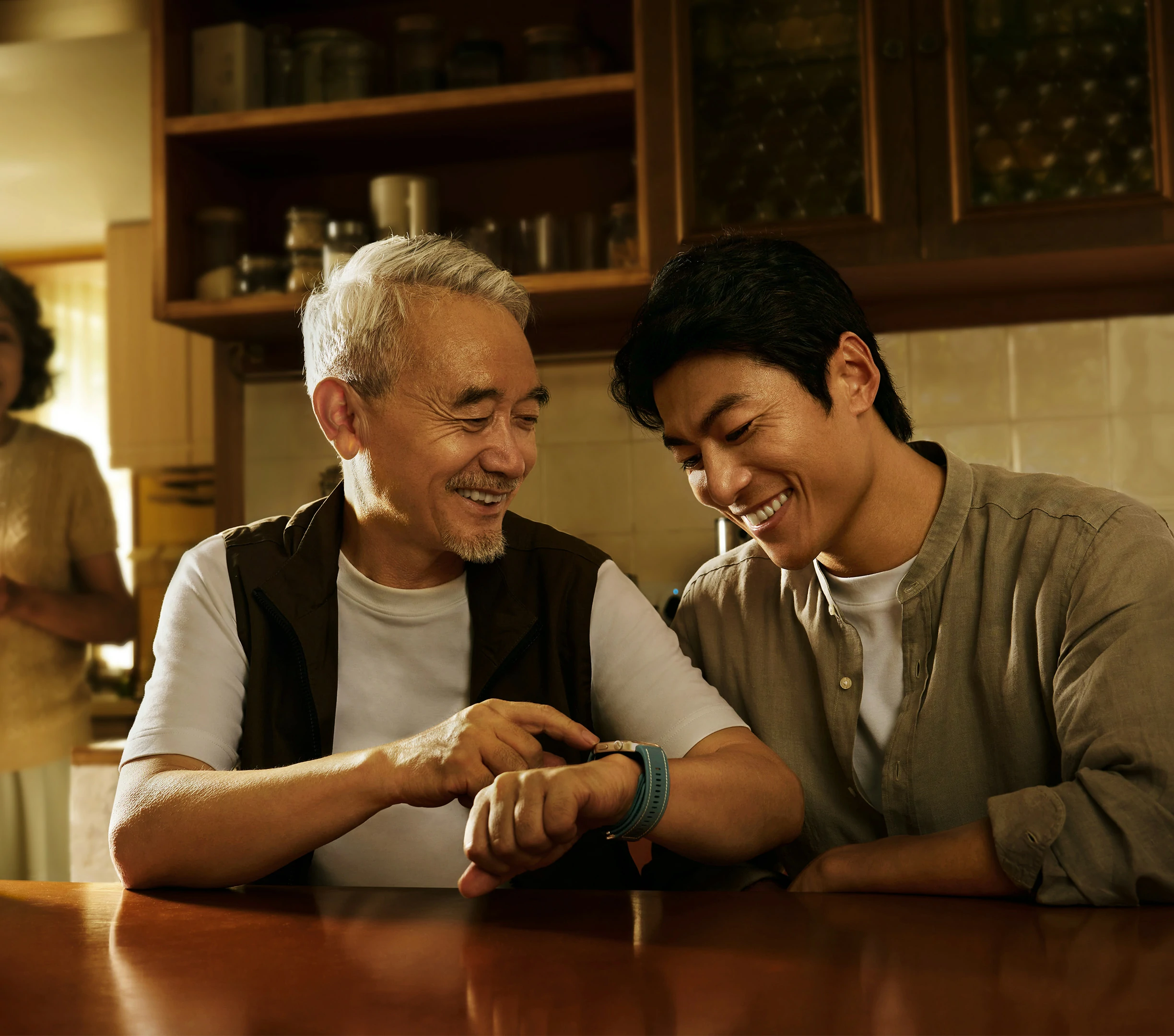 Starszy mężczyzna wskazuje na zegarek na nadgarstku. Młodszy mężczyzna uśmiecha się do zegarka. Kobieta stoi w tle.