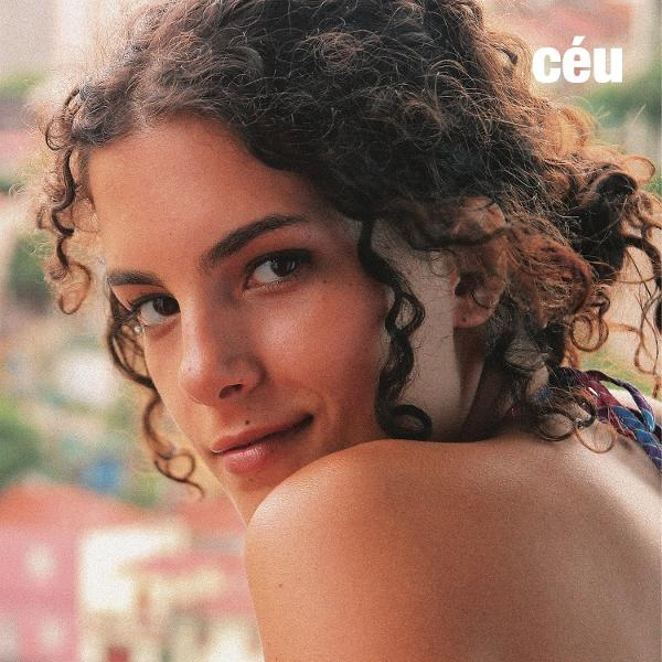 Eine Frau mit lockigem braunem Haar lächelt und schaut über ihre Schulter. Der Hintergrund ist verschwommen mit Andeutungen von Gebäuden.