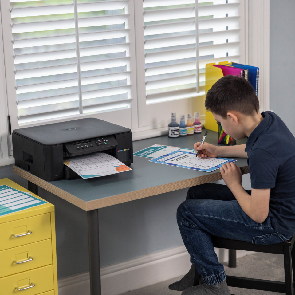 Ein Junge sitzt an einem Schreibtisch und schreibt auf einem Arbeitsblatt. Ein Drucker steht auf dem Schreibtisch und druckt. Im Hintergrund ein Fenster mit Jalousien.