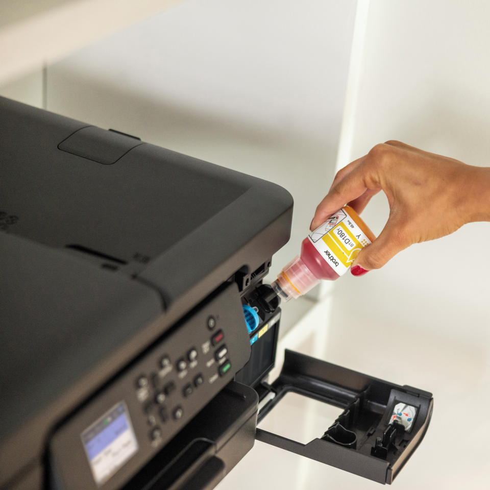 Eine Hand gießt magenta Tinte aus einer gelben Flasche in einen schwarzen Drucker. Der Drucker steht auf einer weißen Oberfläche.