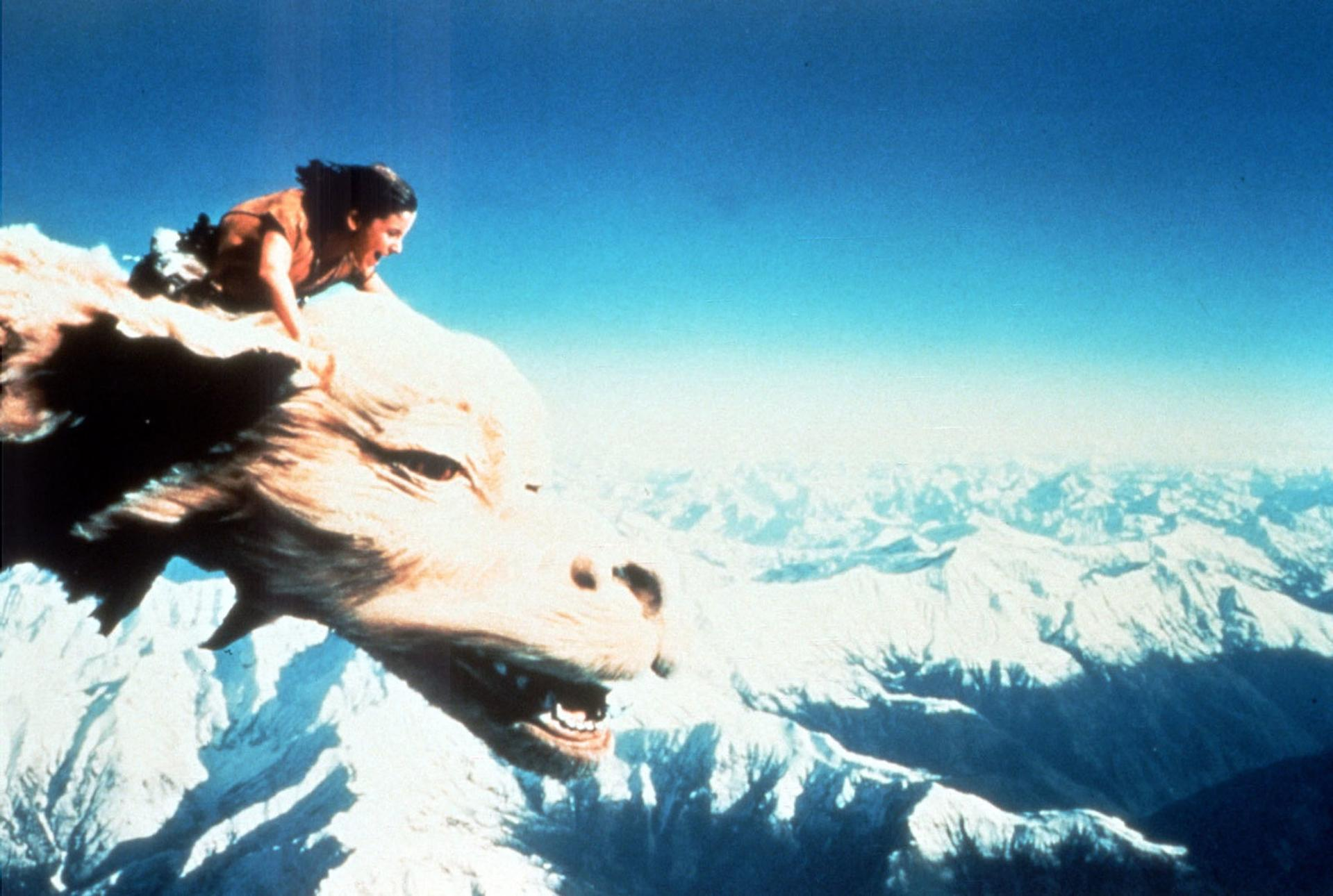 Ein Junge reitet auf einem Drachen über schneebedeckte Berge. Der Drache ist hell, der Himmel blau mit hellem Horizont.