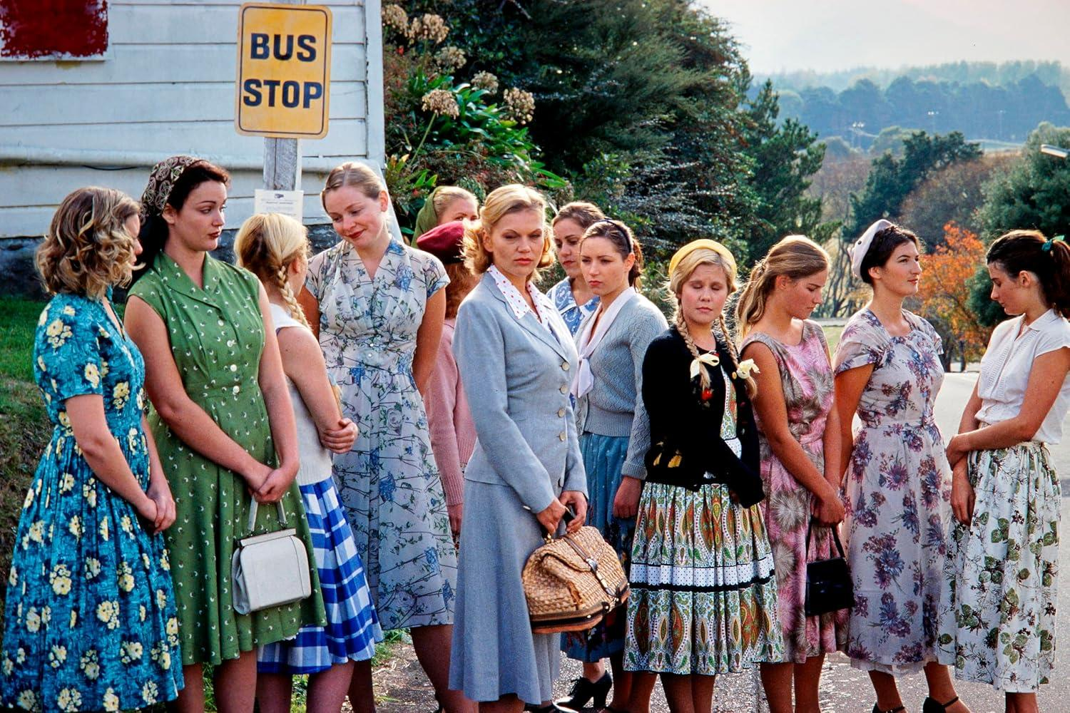 Eine Gruppe von Frauen in Vintage-Kleidern steht an einer Bushaltestelle. Ein Schild zeigt 'Bushaltestelle'. Einige halten Handtaschen. Die Szene ist im Freien.