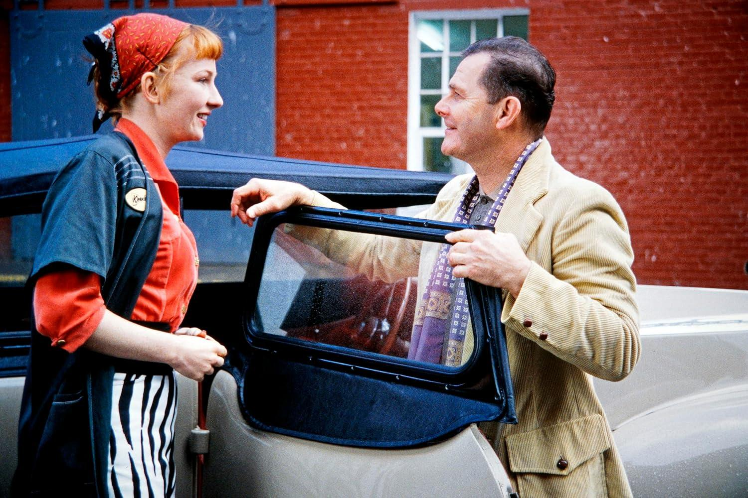 Eine Frau mit rotem Bandana und ein Mann mit beiger Jacke unterhalten sich an einem Auto. Im Hintergrund ein rotes Backsteingebäude.