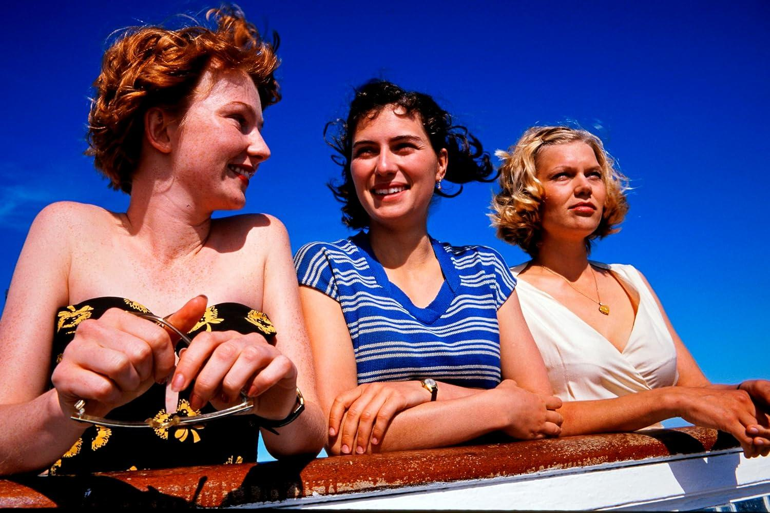 Drei Frauen auf einem Bootsdeck, blauer Himmel im Hintergrund. Eine mit rotem Haar, einem Oberteil mit Blumenmuster und Sonnenbrille, die anderen lächeln.