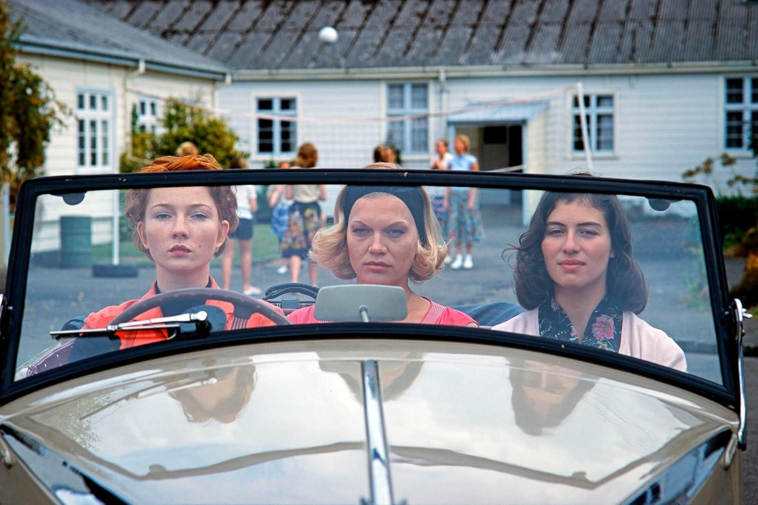Drei Frauen sitzen in einem Oldtimer. Die Fahrerin hat rotes Haar, die mittlere Frau blondes Haar, die dritte dunkles Haar.