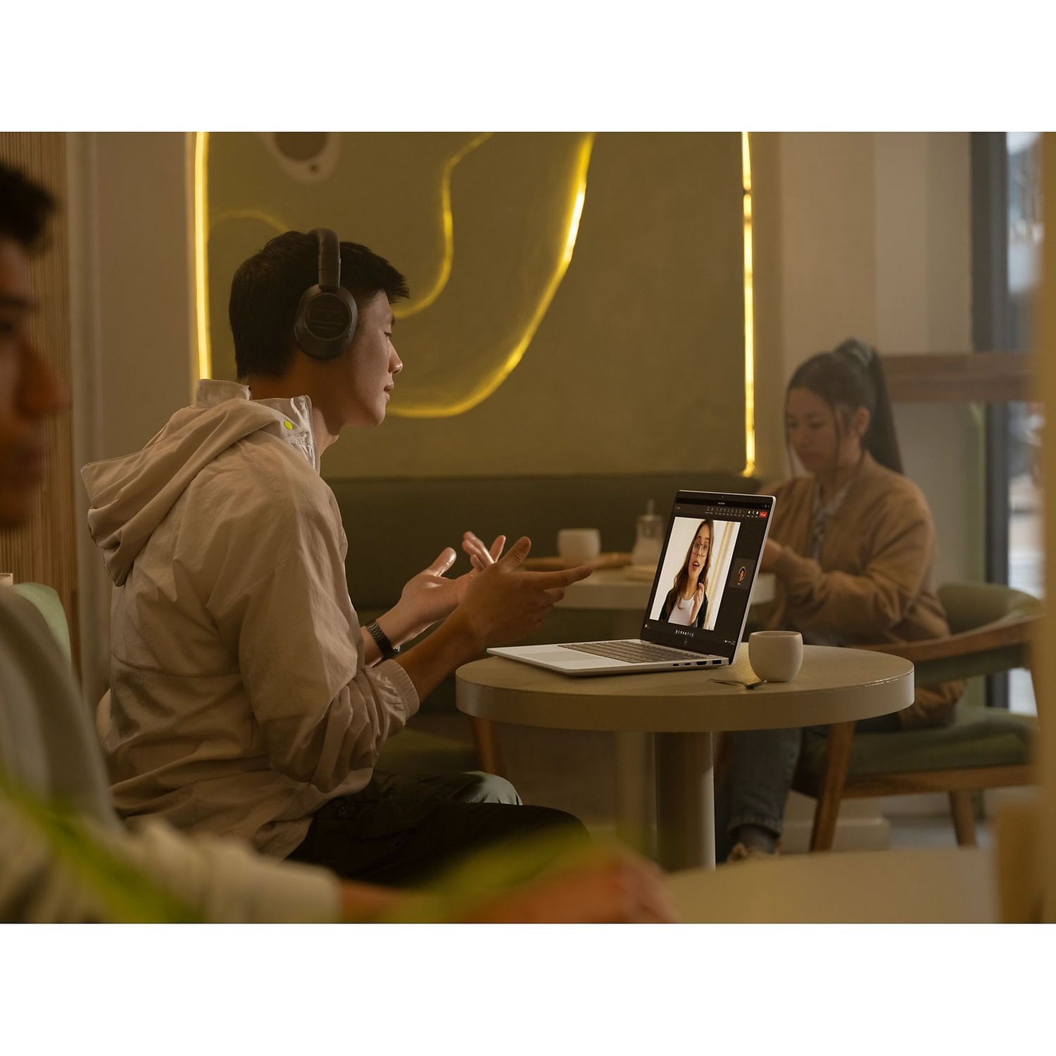 A man with headphones gestures while on a video call. A woman is on screen. Another woman sits in the background, and another man in the foreground.