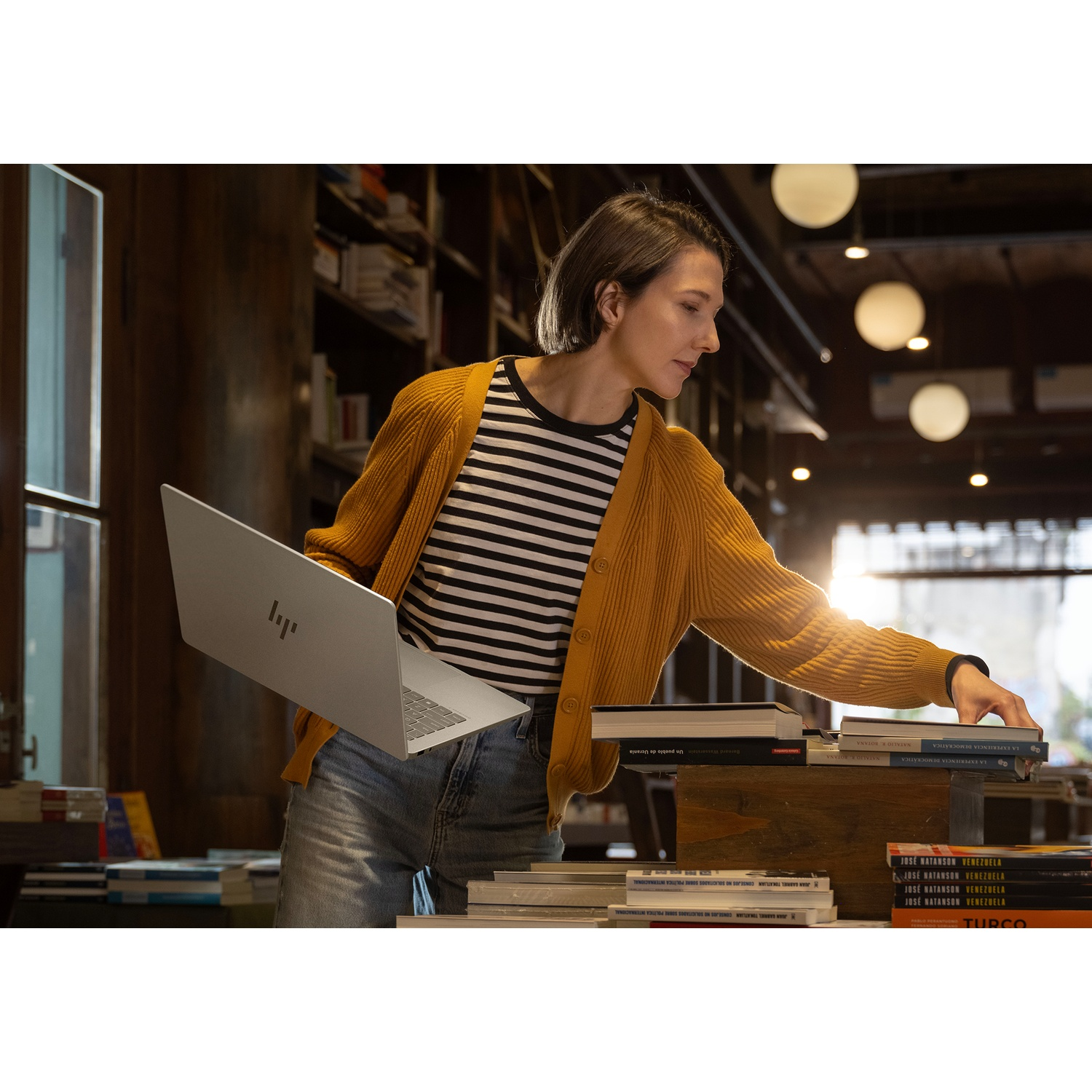 A woman in a yellow cardigan and striped shirt in a library, holding a laptop and selecting a book. Bookshelves are in background.