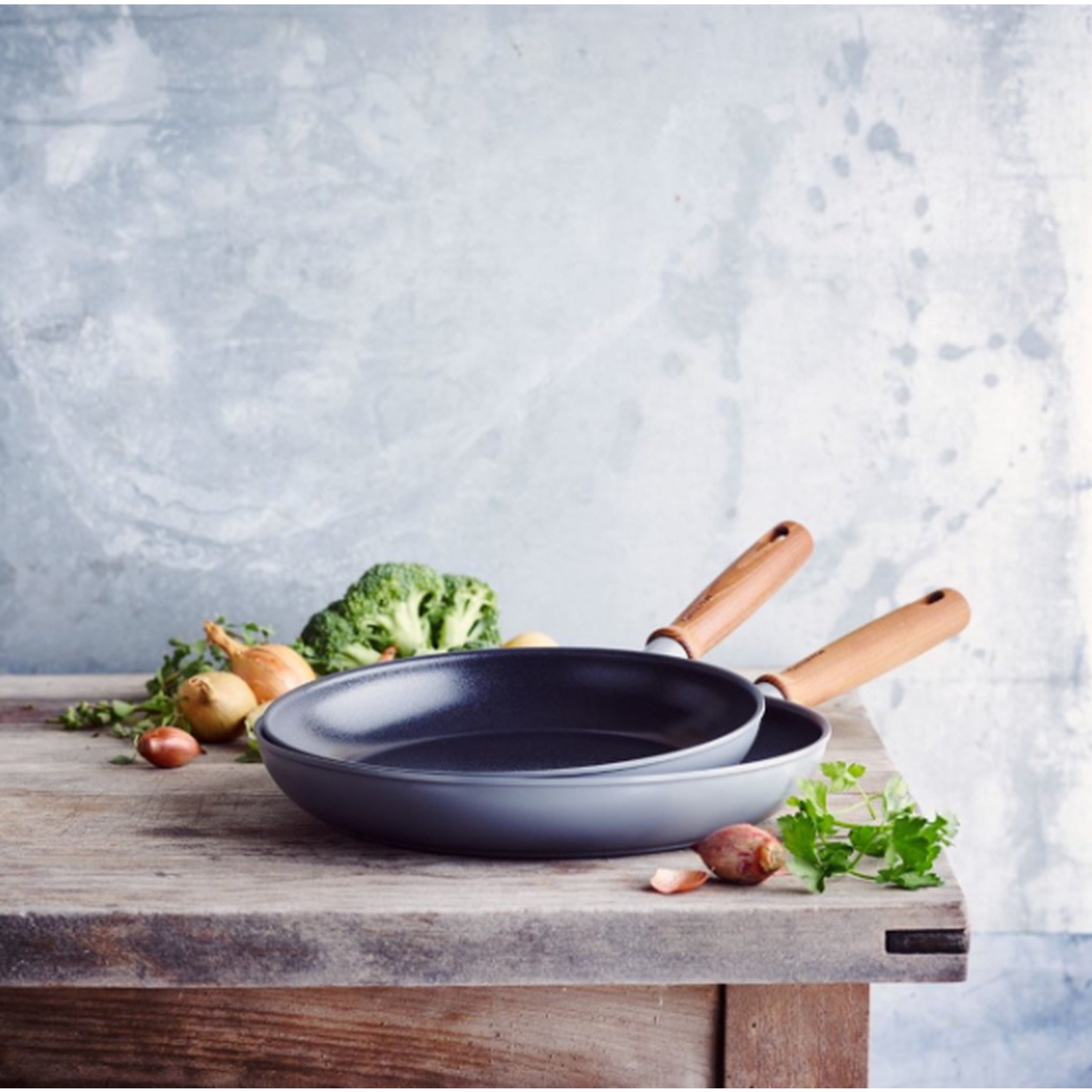 Two pans on a wooden table with vegetables like broccoli, onions, and garlic. The background is a light blue wall.