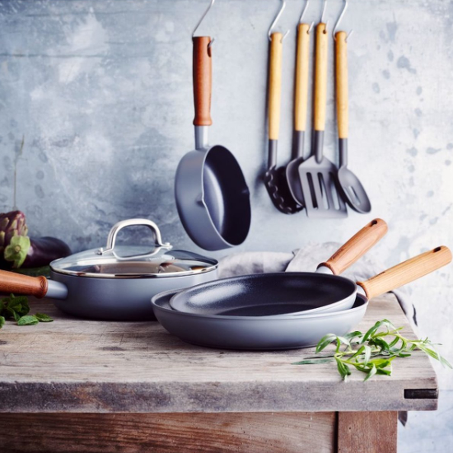 Kitchen scene. Pans, utensils, and herbs are on a wooden table. Background is a metallic wall. Some items are hanging.