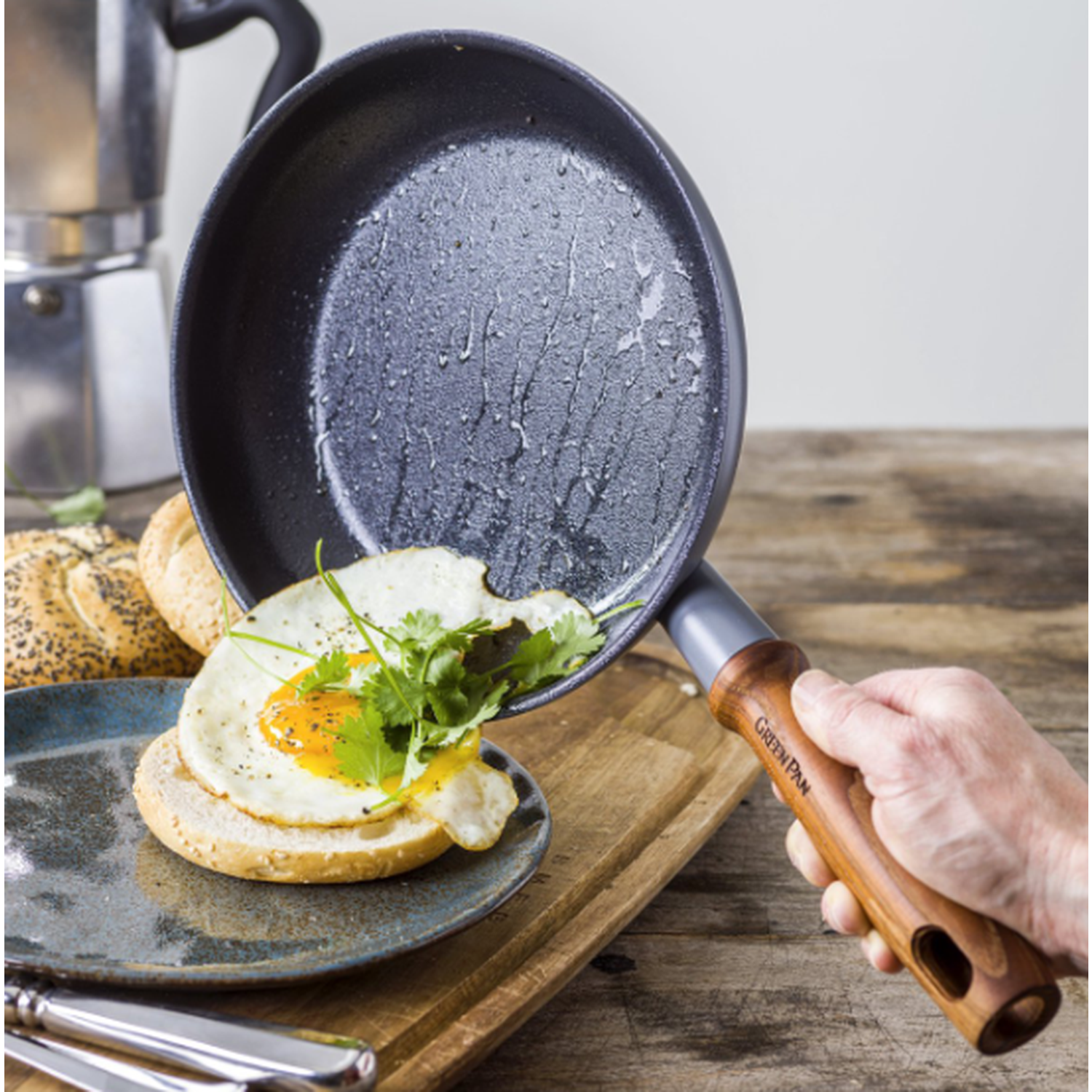 A fried egg with herbs on a bun is being placed on a plate. A pan with oil is tilted. A hand holds the pan's wooden handle.